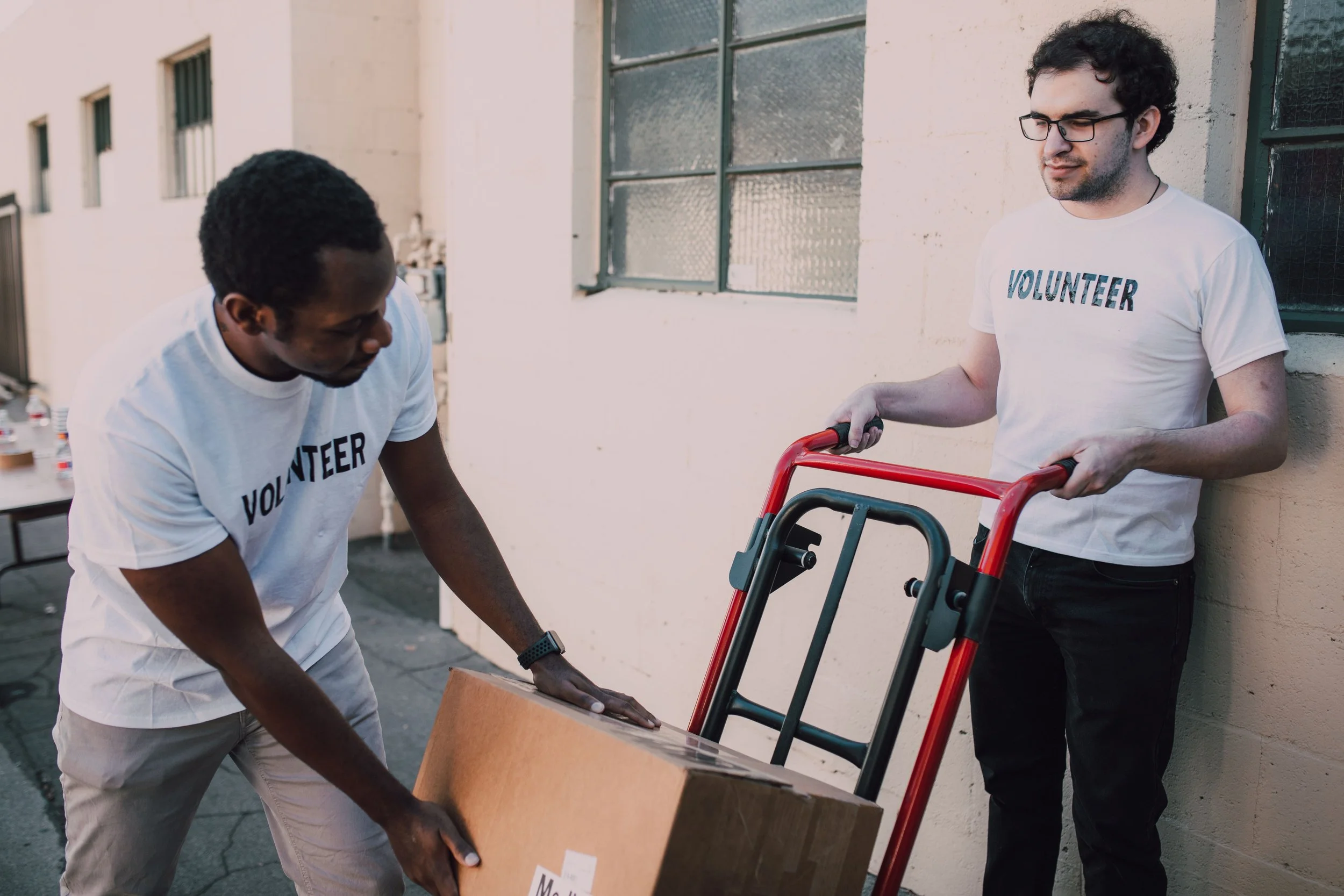 Two men wearing white t-shirts with the word 'VOLUNTEER' on them are working together outdoors. One man with glasses is holding a red hand truck, and the other man is lifting a cardboard box.