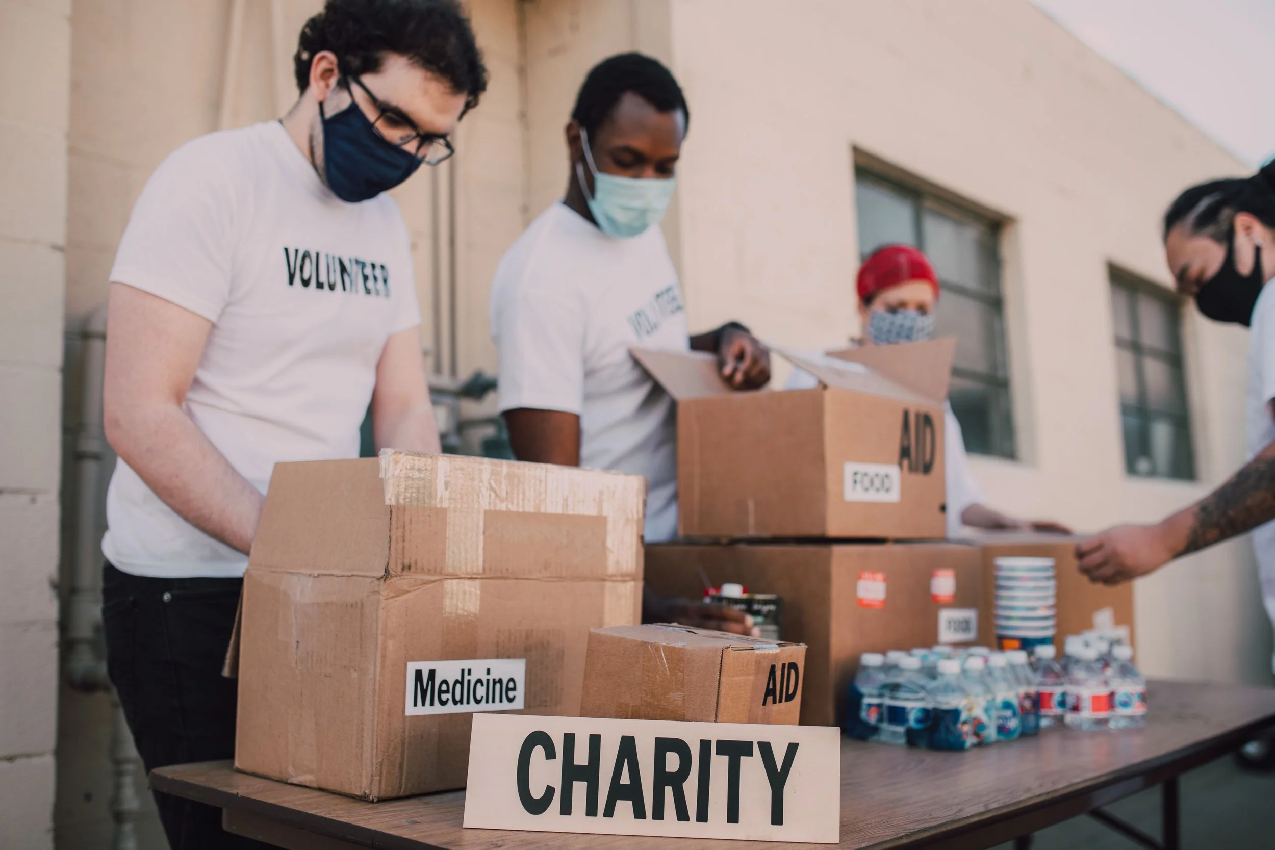 Group of diverse volunteers wearing masks, packing boxes labeled 'Medicine', 'AID', and 'Charity' outside a building for a charity drive.