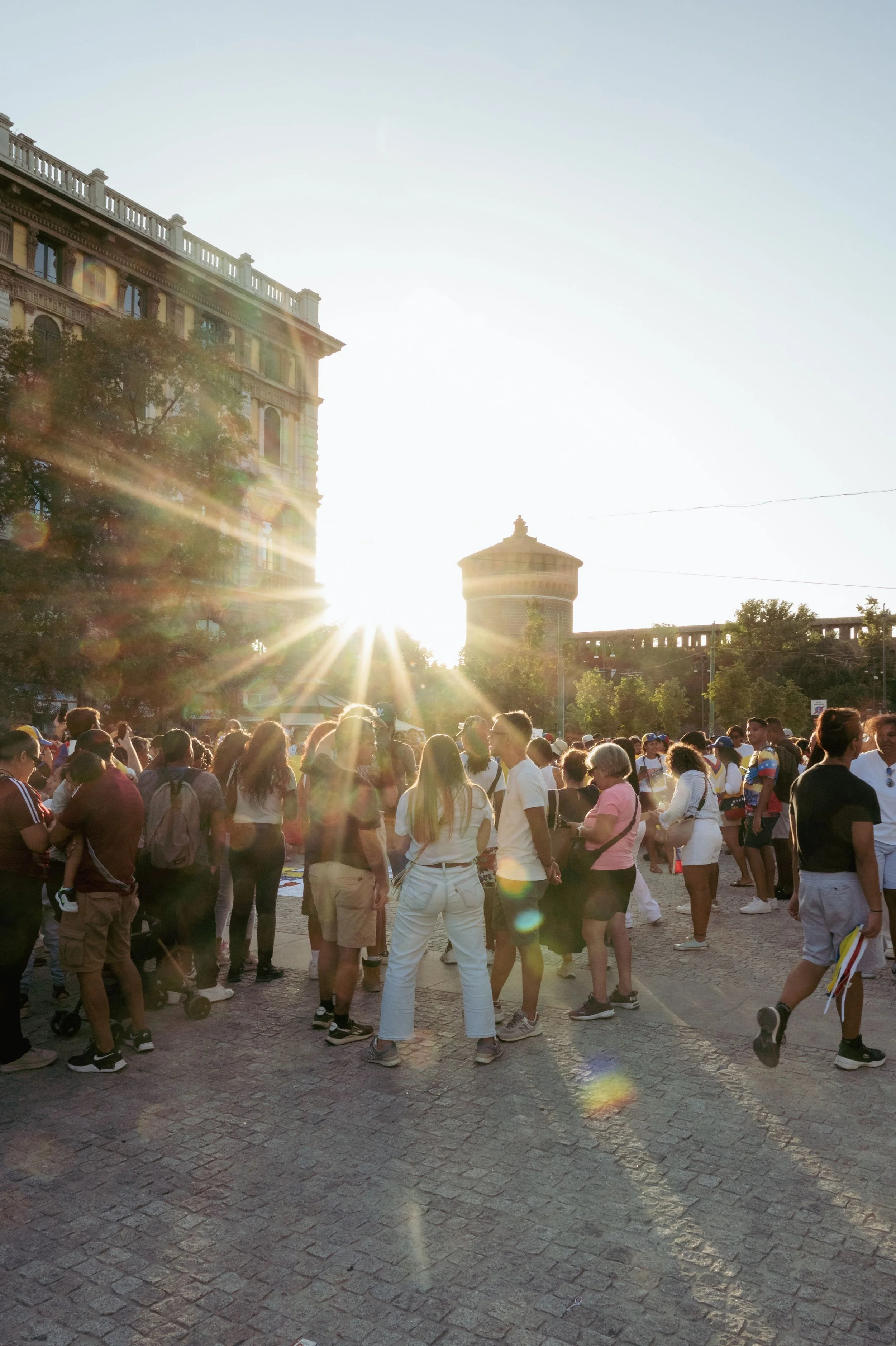 Crowd of people outdoors, looking towards the sunlight, in front of historic buildings and a water tower, during sunset or sunrise.