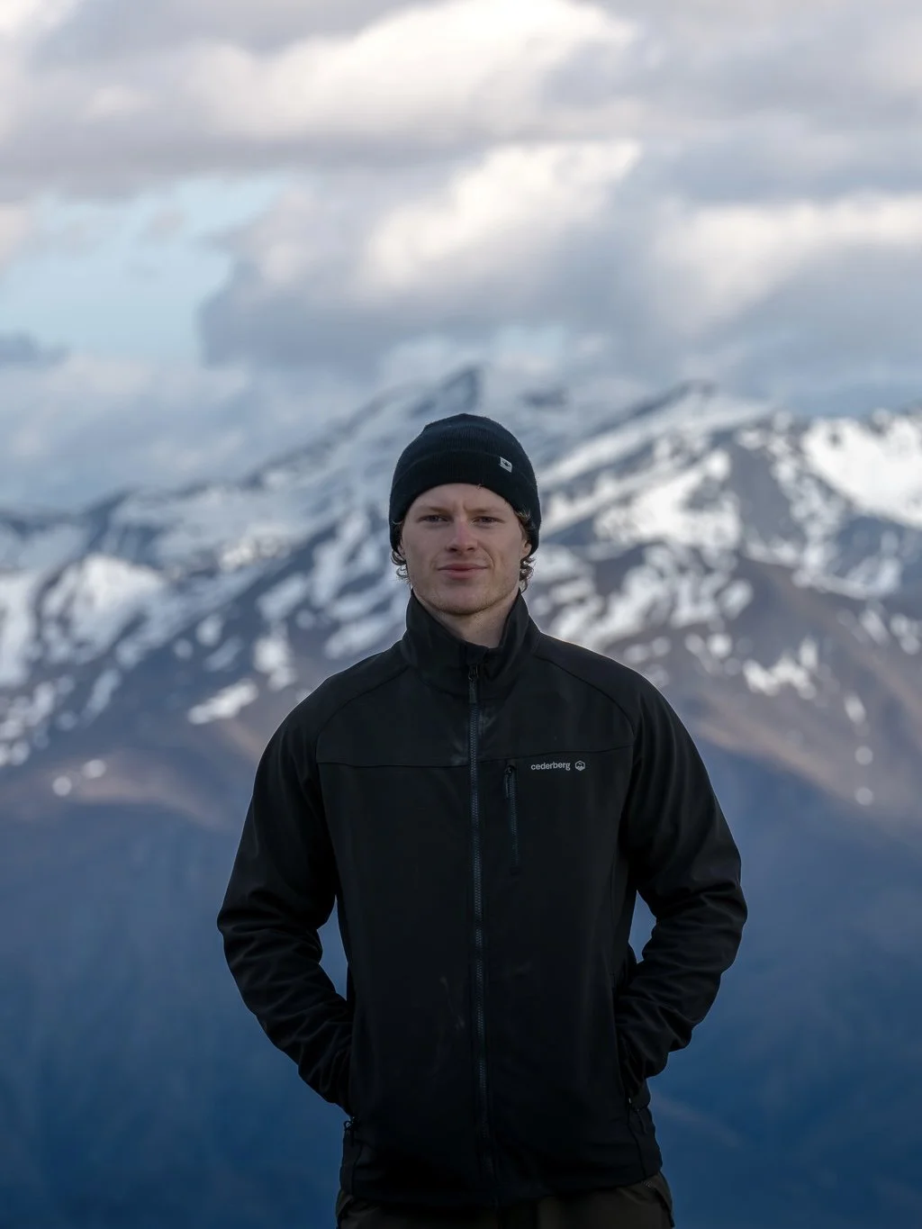 Young man standing outdoors with snow-capped mountains and cloudy sky in the background.