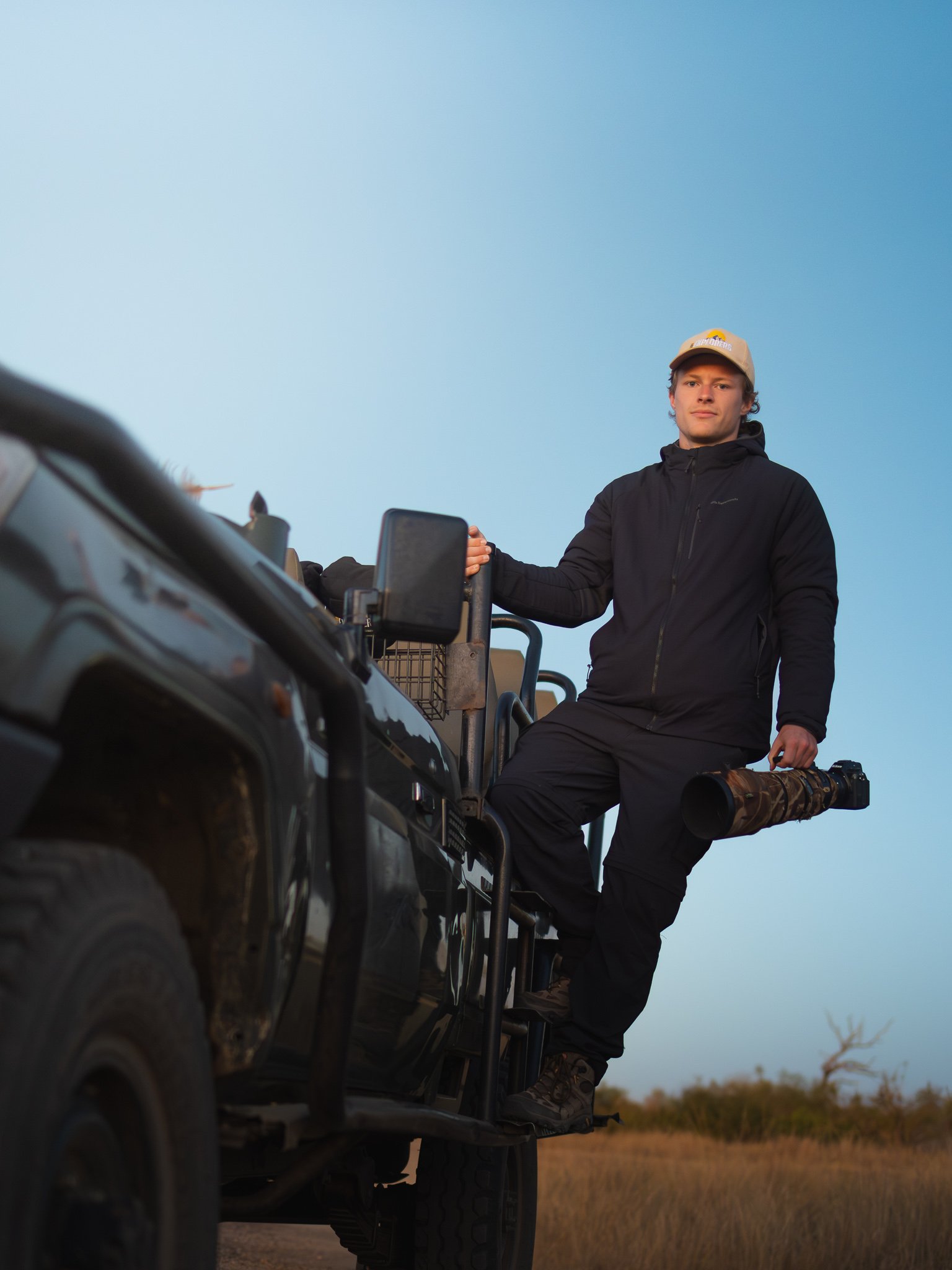 A young man standing on the side of a vehicle, outdoors during the day with a clear blue sky in the background, holding a camera with a large telephoto lens.