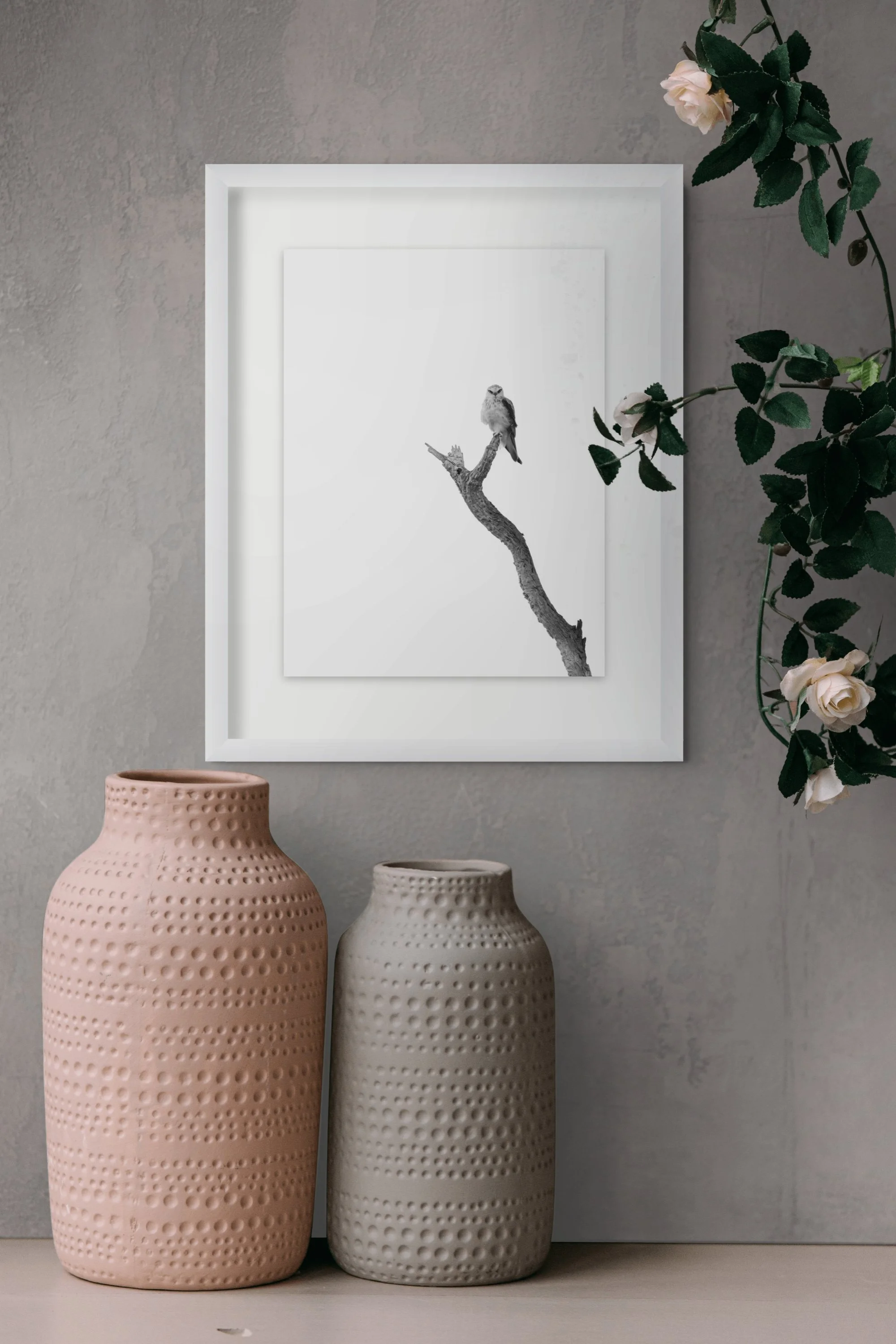 Two tall ceramic vases, one pink and one gray, with a textured dotted pattern, sit side by side. Behind them is a framed black and white photograph of a bird perched on a branch. To the right, green leafy plant with white flowers extends into the frame.