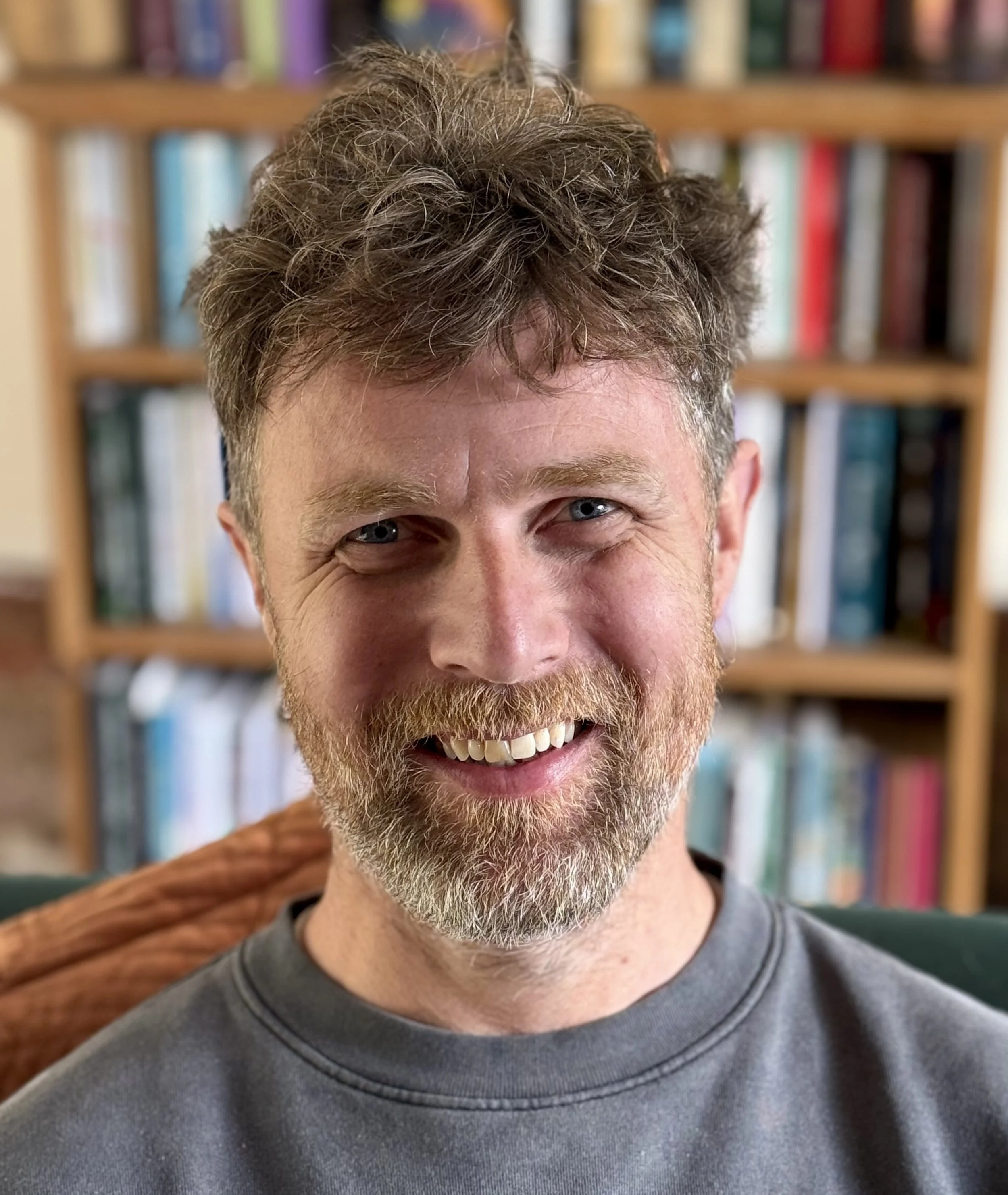 Close-up of a smiling man with gray and brown curly hair, blue eyes, a beard, wearing a gray shirt, with a wooden bookshelf filled with books in the background.