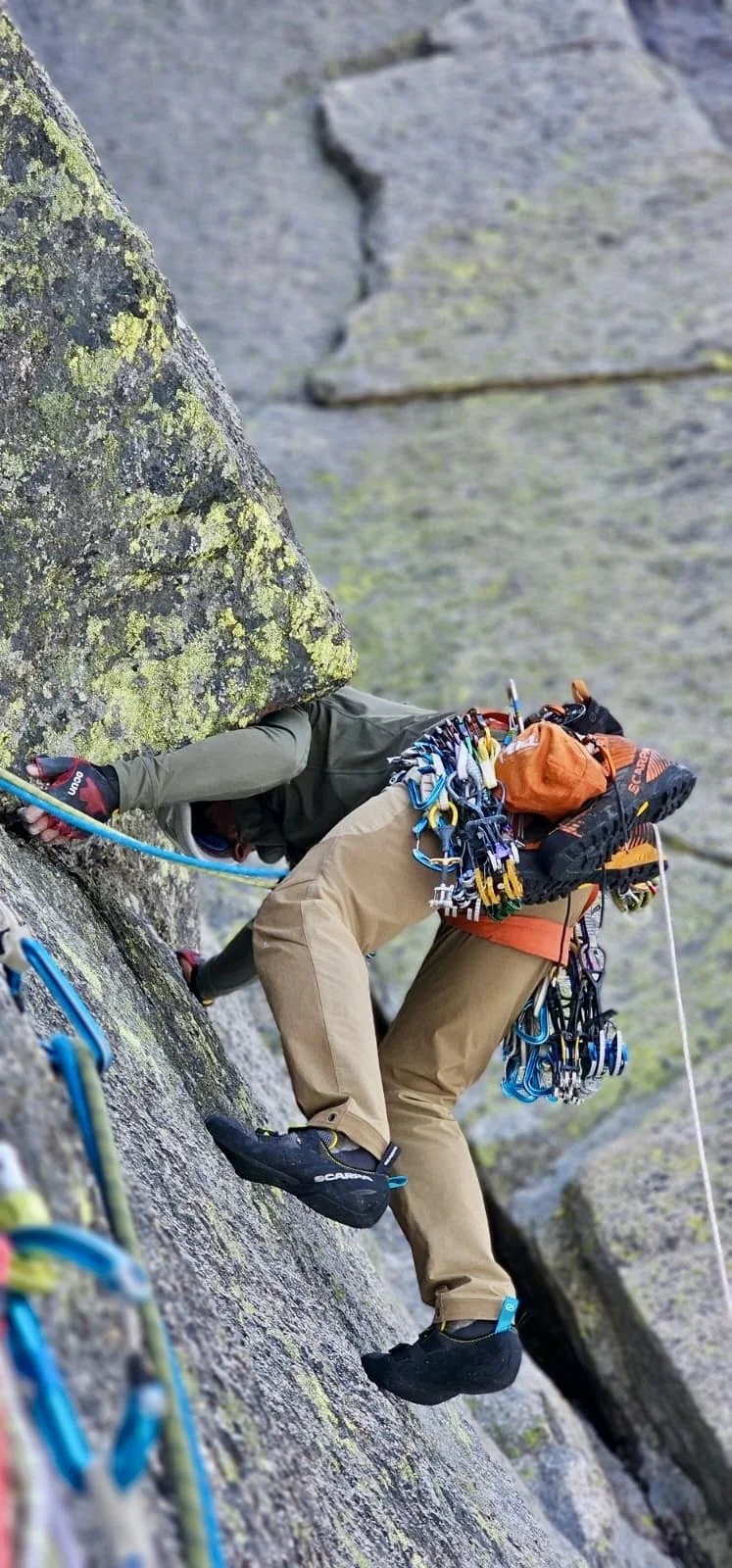 Alpiniste grimpeant une falaise rocheuse, équipé de matériel d'escalade, portant un sac et un casque, en train de progressant sur une paroi rocheuse verticale.