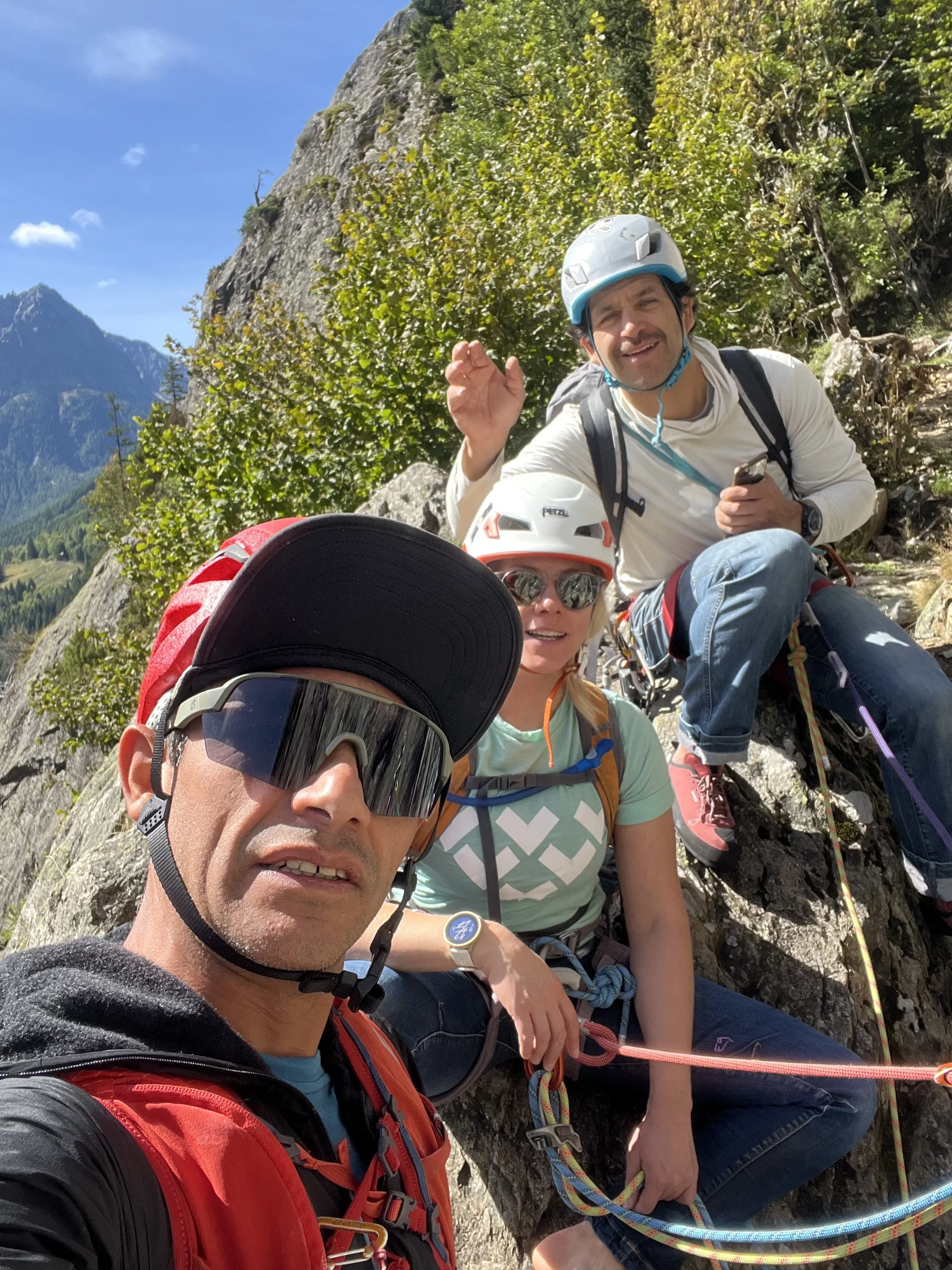 Groupe de cinq personnes en pleine escalade en montagne, avec des casques et de l'équipement d'escalade, dans un paysage naturel avec des montagnes et des arbres.