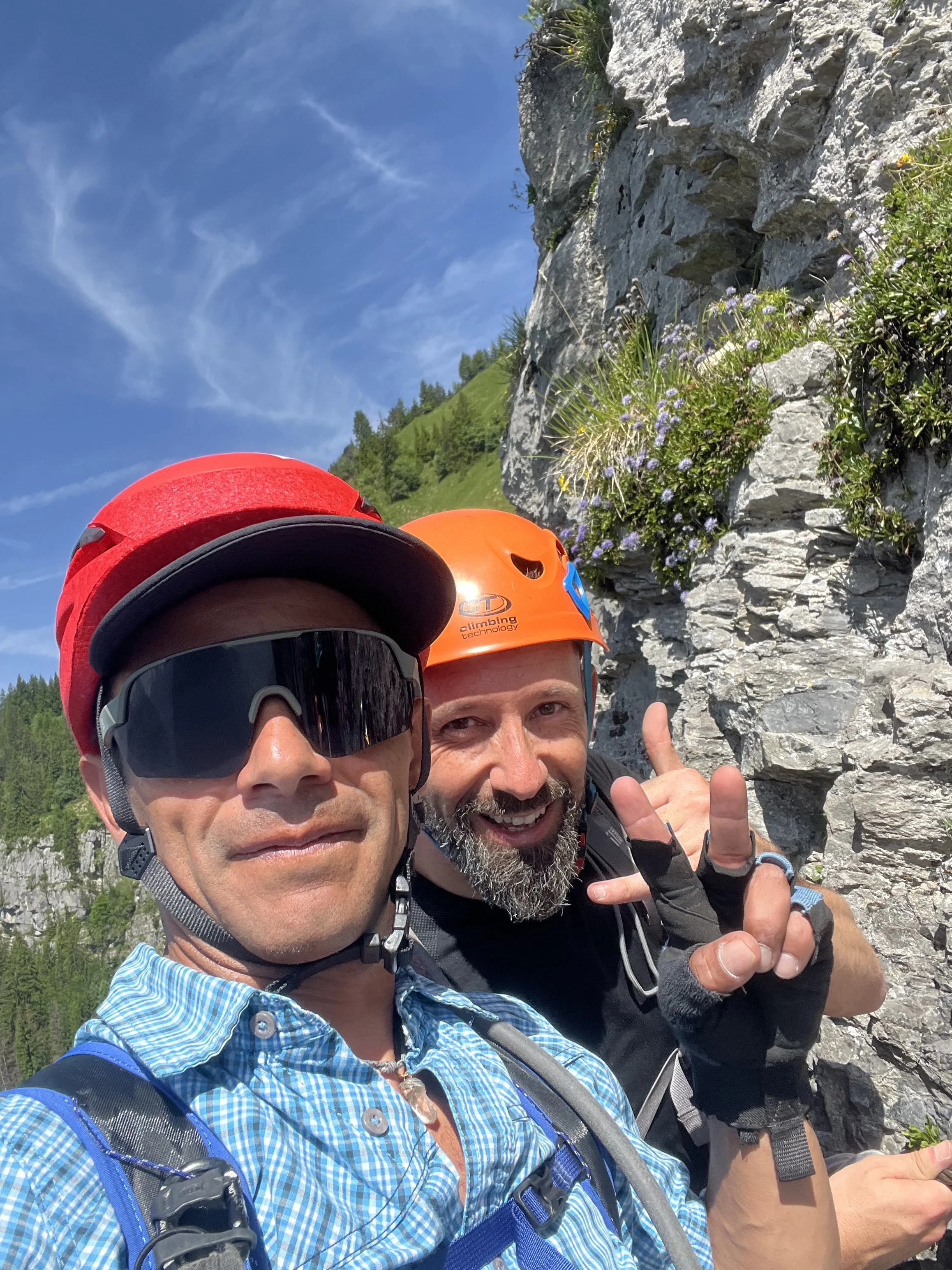 Deux hommes en équipement d'escalade posant pour une photo en montagne, avec un ciel bleu et des formations rocheuses en arrière-plan, l'un d'eux fait un signe de paix.
