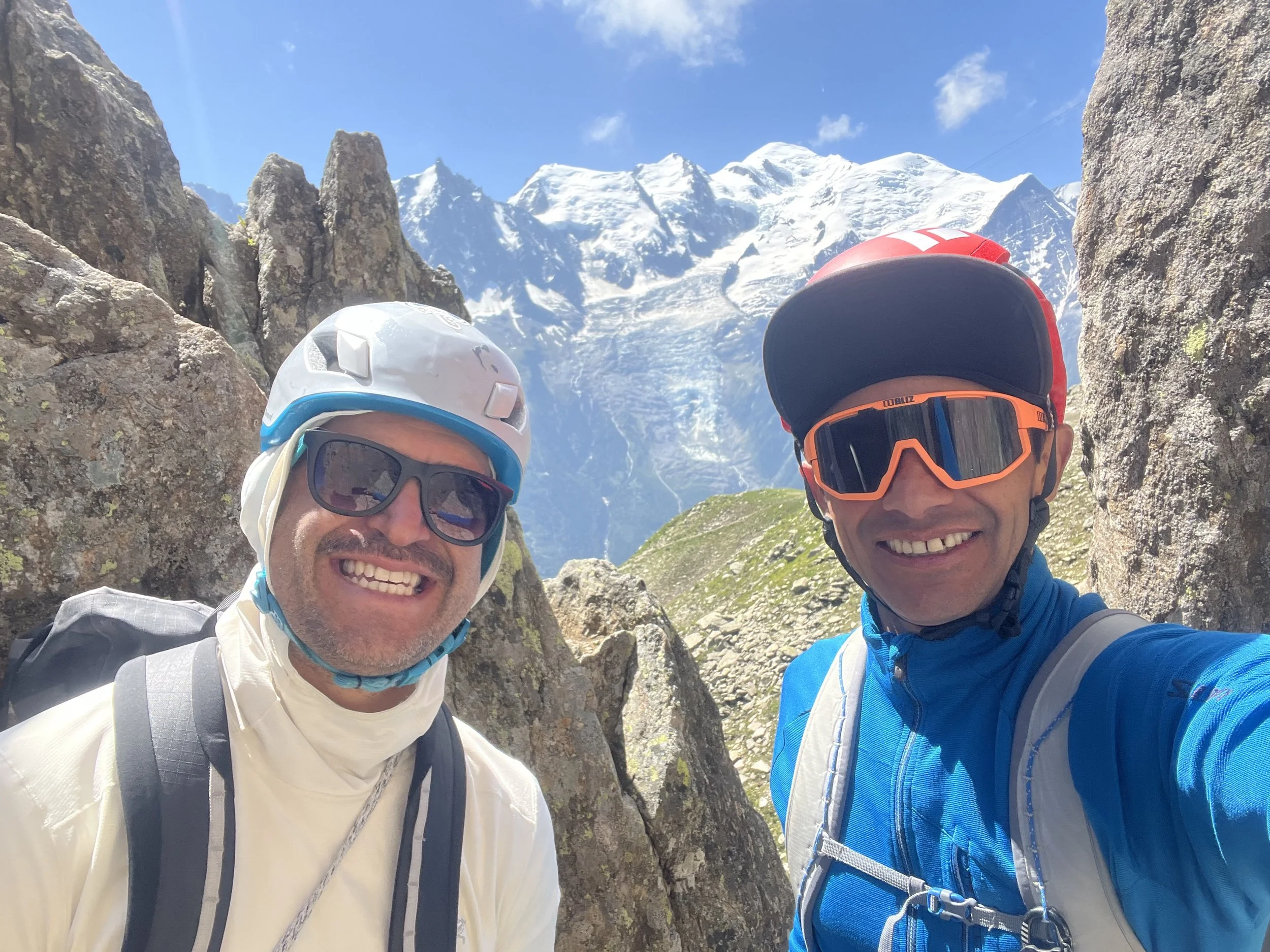 Deux hommes portant des casques de montagne, lunettes de soleil et tenues de randonnée, prenant un selfie dans un paysage alpin avec des montagnes enneigées en arrière-plan.
