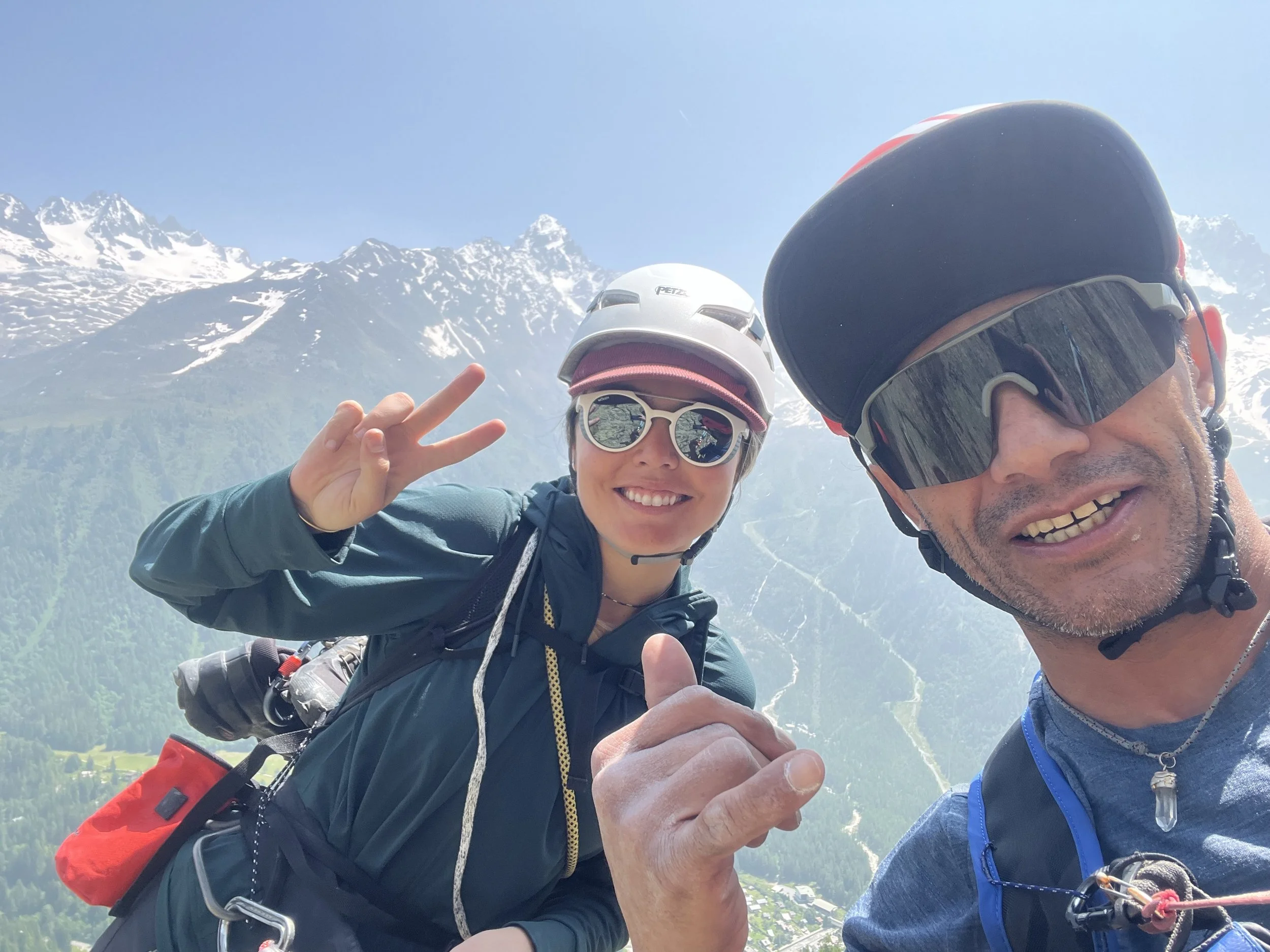 Deux personnes portant des casques de vélo, avec des montagnes enneigées en arrière-plan, prennent un selfie en montagne, faisant un signe de paix et souriant.
