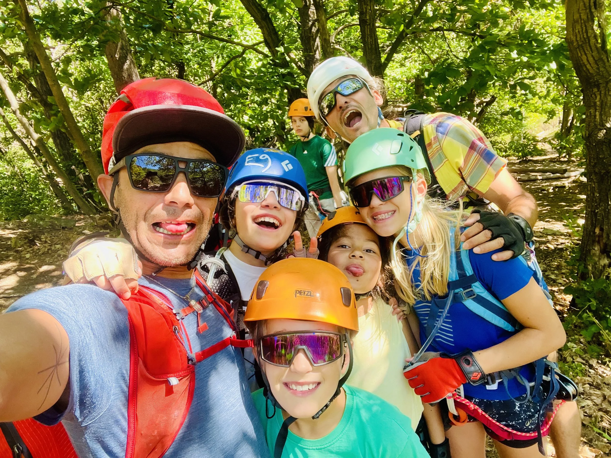 Groupe de six personnes équipées d'armures de vélo, casques et lunettes de soleil, posant pour une photo dans une forêt dense, souriant et montrant des gestes de paix.