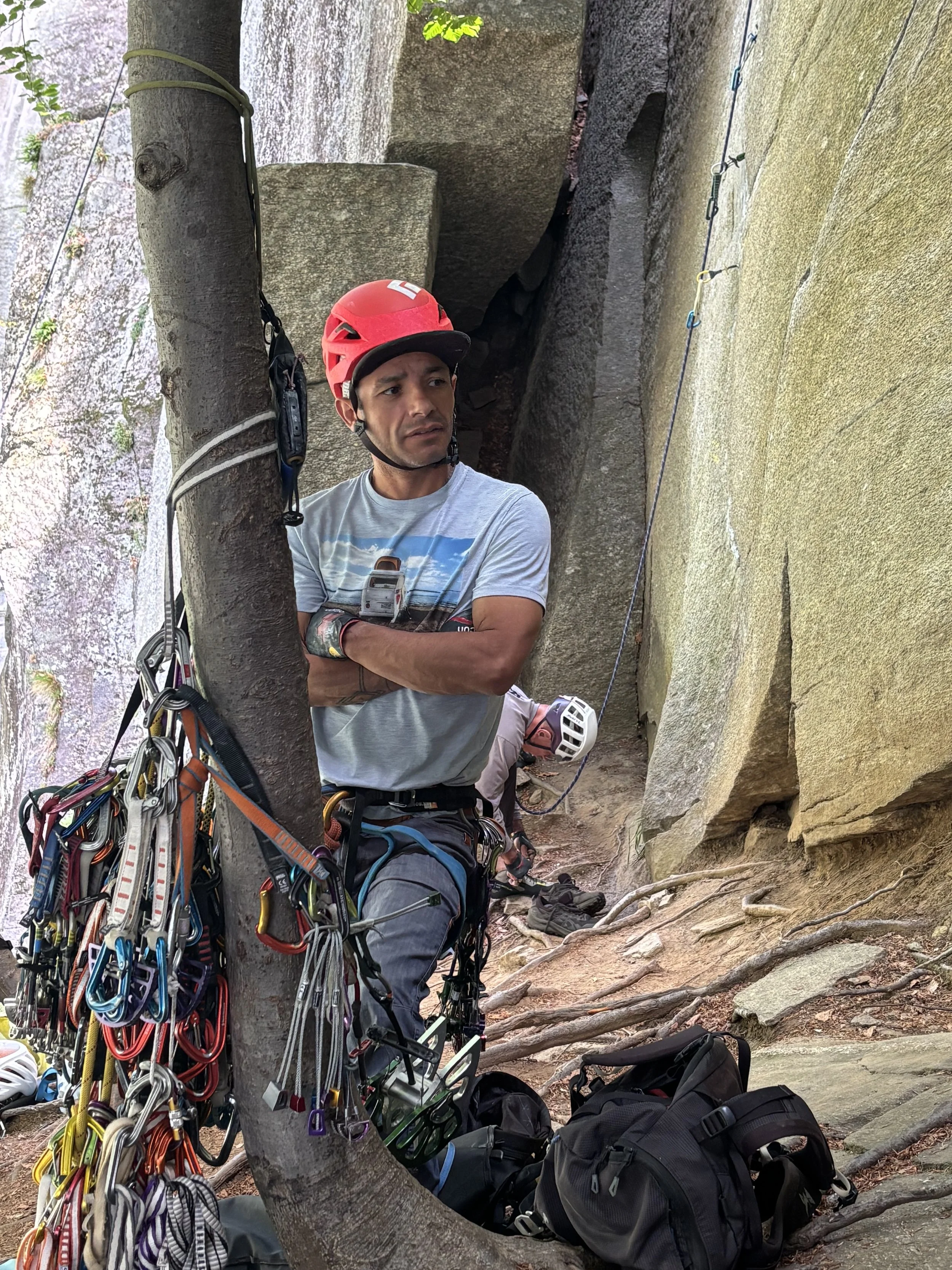 Un homme avec un casque rouge et une chemise graphique, qui se prépare à faire de l'escalade ou de la rappel, entouré de matériel d'escalade, devant une paroi rocheuse.