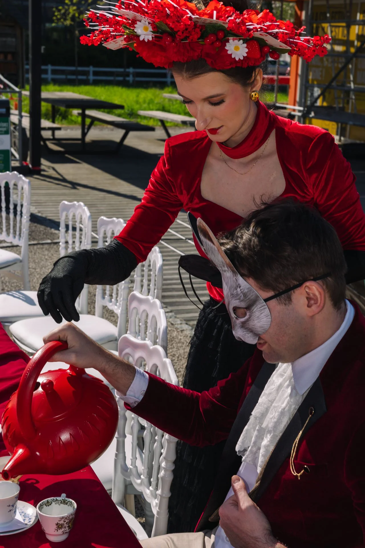 Une femme portant un chapeau de fleurs rouges et une robe rouge sert du thé à un homme portant un masque de lapin lors d'une fête en plein air.