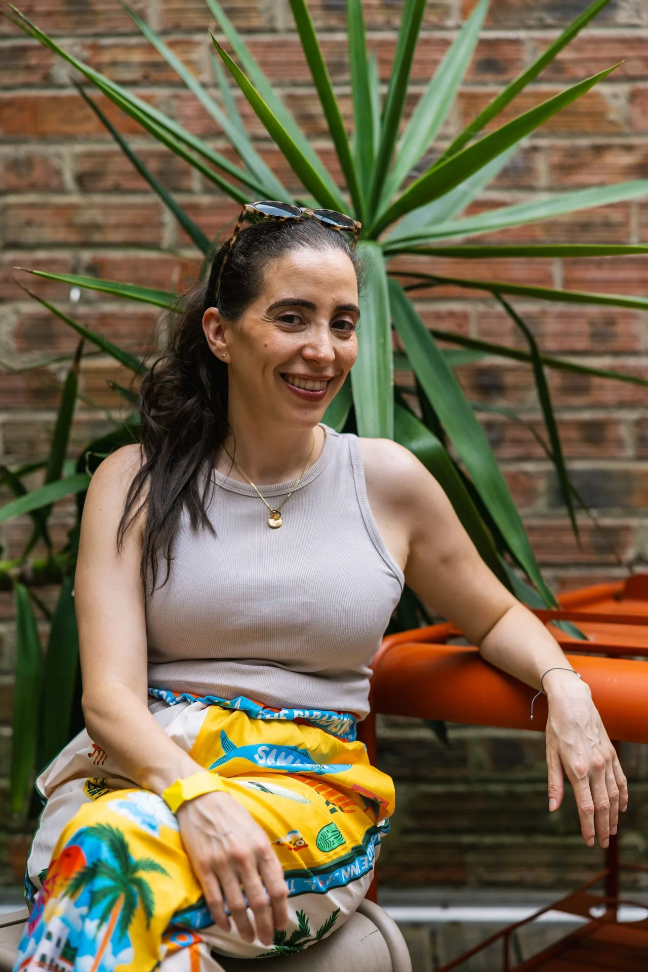 Une femme souriante portant des lunettes de soleil sur sa tête, assise devant un mur en briques et une grande plante verte, vêtue d'un haut gris et d'un pantalon coloré avec des motifs de fleurs et de palmiers.