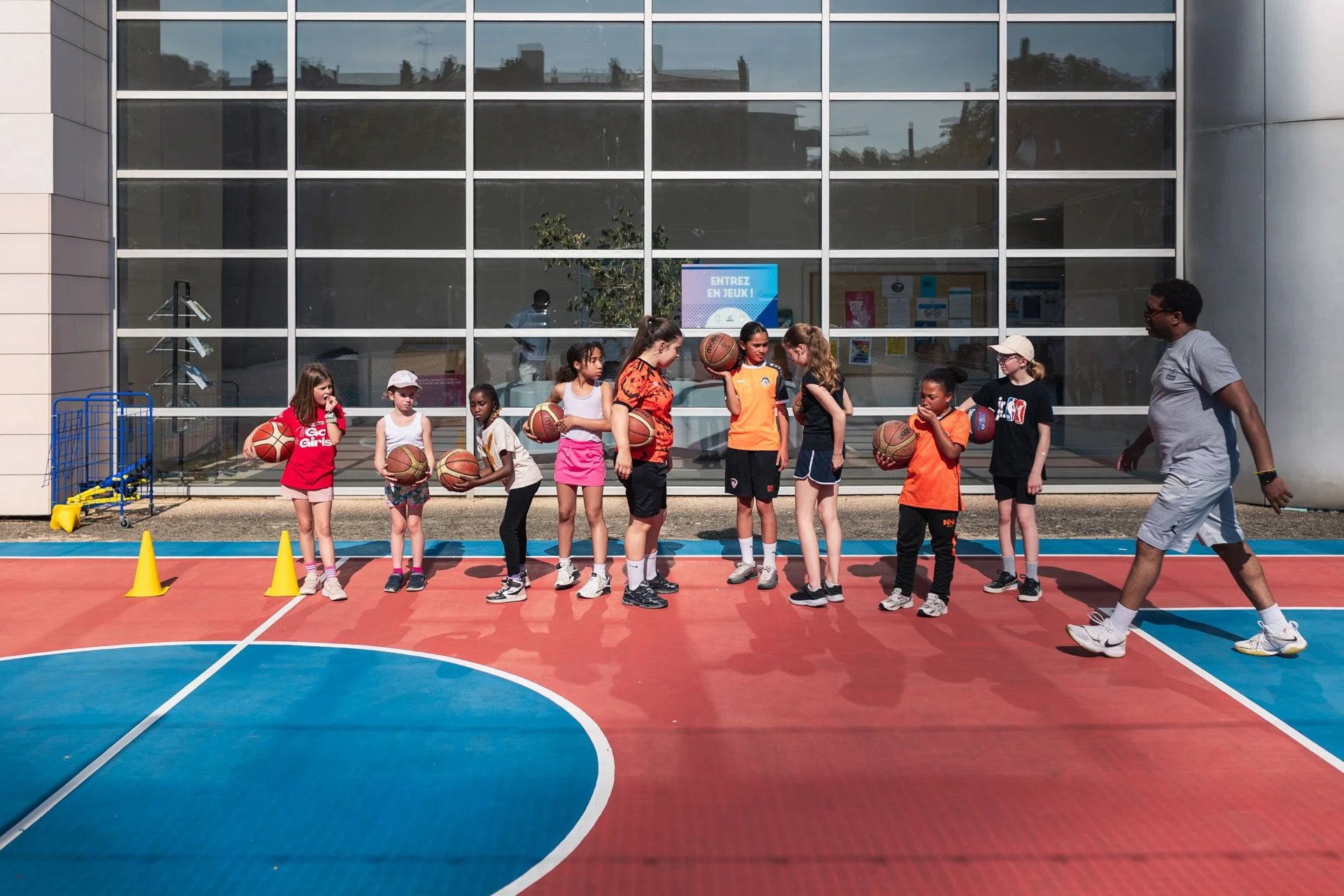 Groupe d'enfants jouant au basketball sous la supervision d'un entraîneur sur un terrain extérieur.