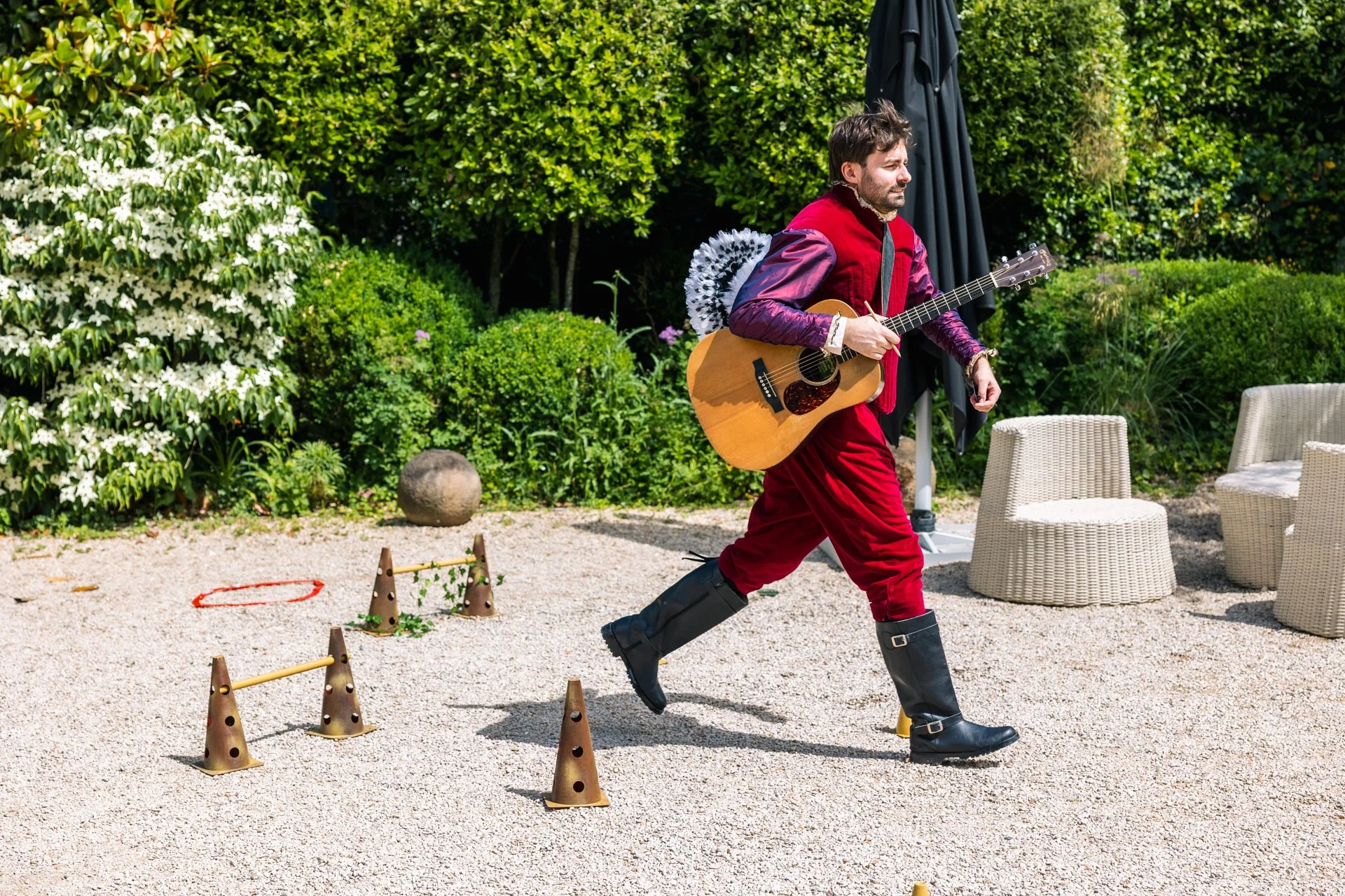 Un homme déguisé en piéton romain avec une guitare, marchant dans un jardin en été, avec des plantes et des chaises en rotin, des cônes de signalisation et des cerceaux sur le sol.