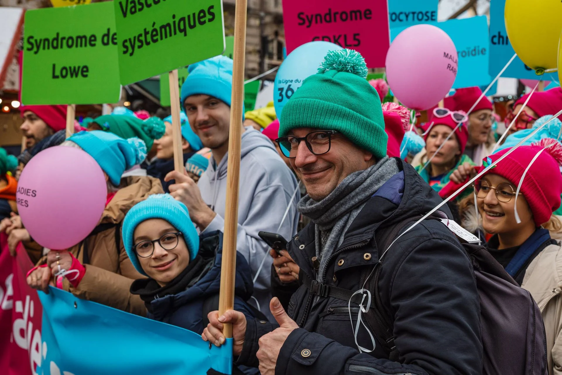 Groupe de personnes participant à une marche ou une manifestation, portant des chapeaux colorés et tenant des ballons, avec des pancartes en français mentionnant des syndromes génétiques et des lignes de métro.
