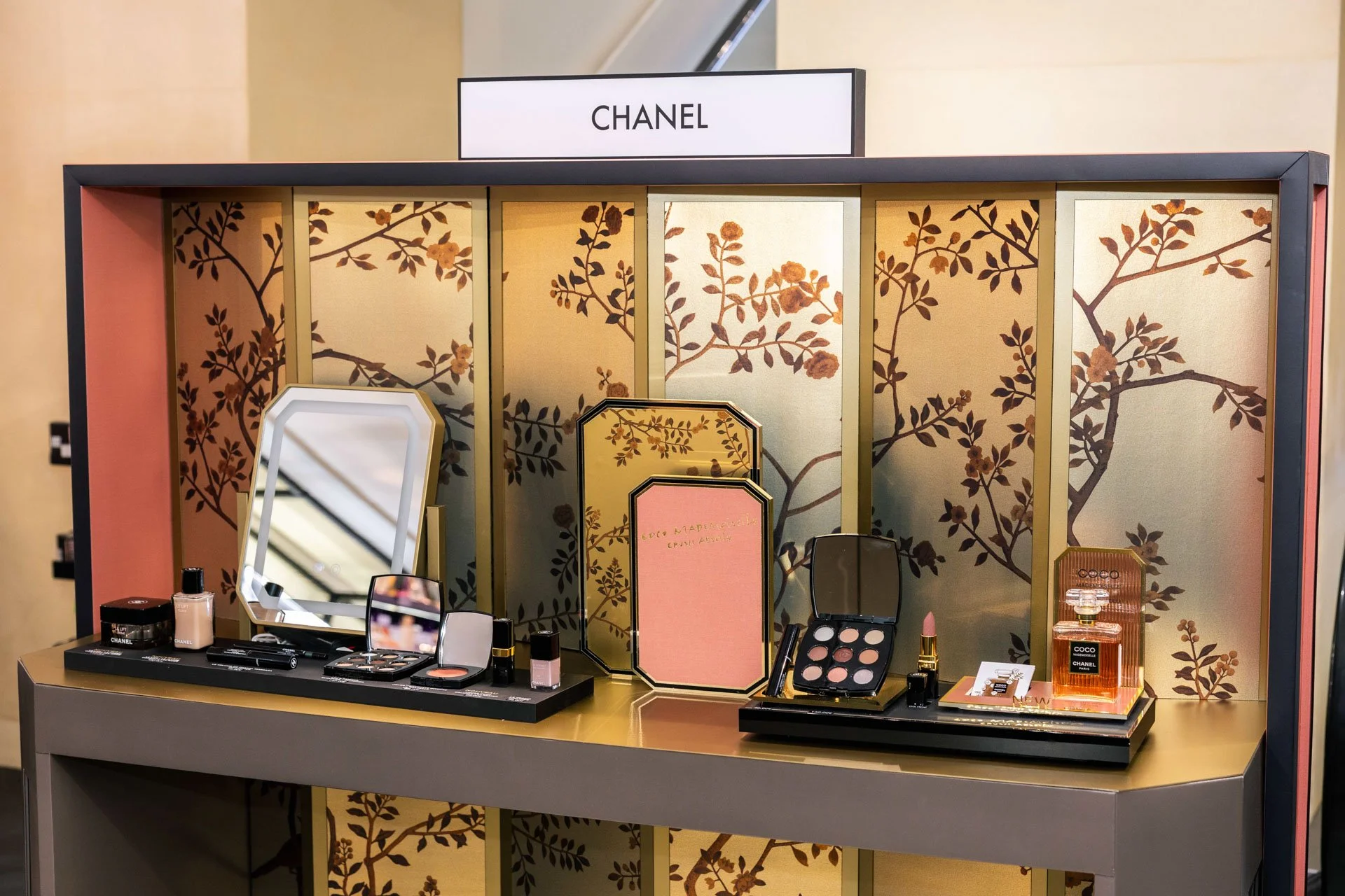 Présentation de produits cosmétiques Chanel, incluant maquillage, parfums et soins, sur une table, avec un fond décoratif floral doré.