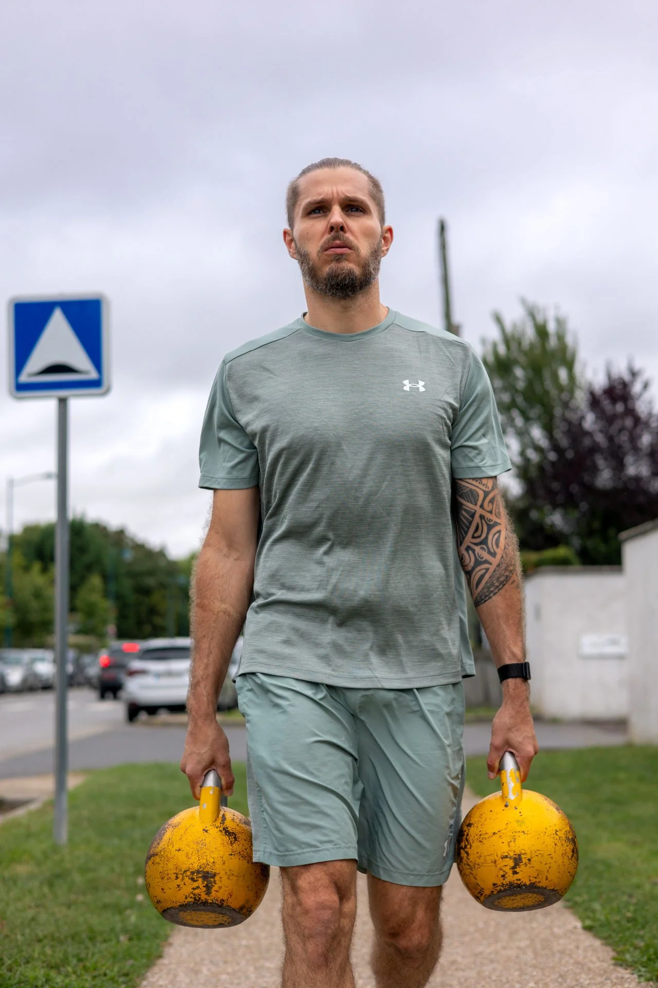 Un homme musclé fait de l'exercice en marchant avec deux kettlebells jaunes dans ses mains sur un trottoir en ville, sous un ciel nuageux.