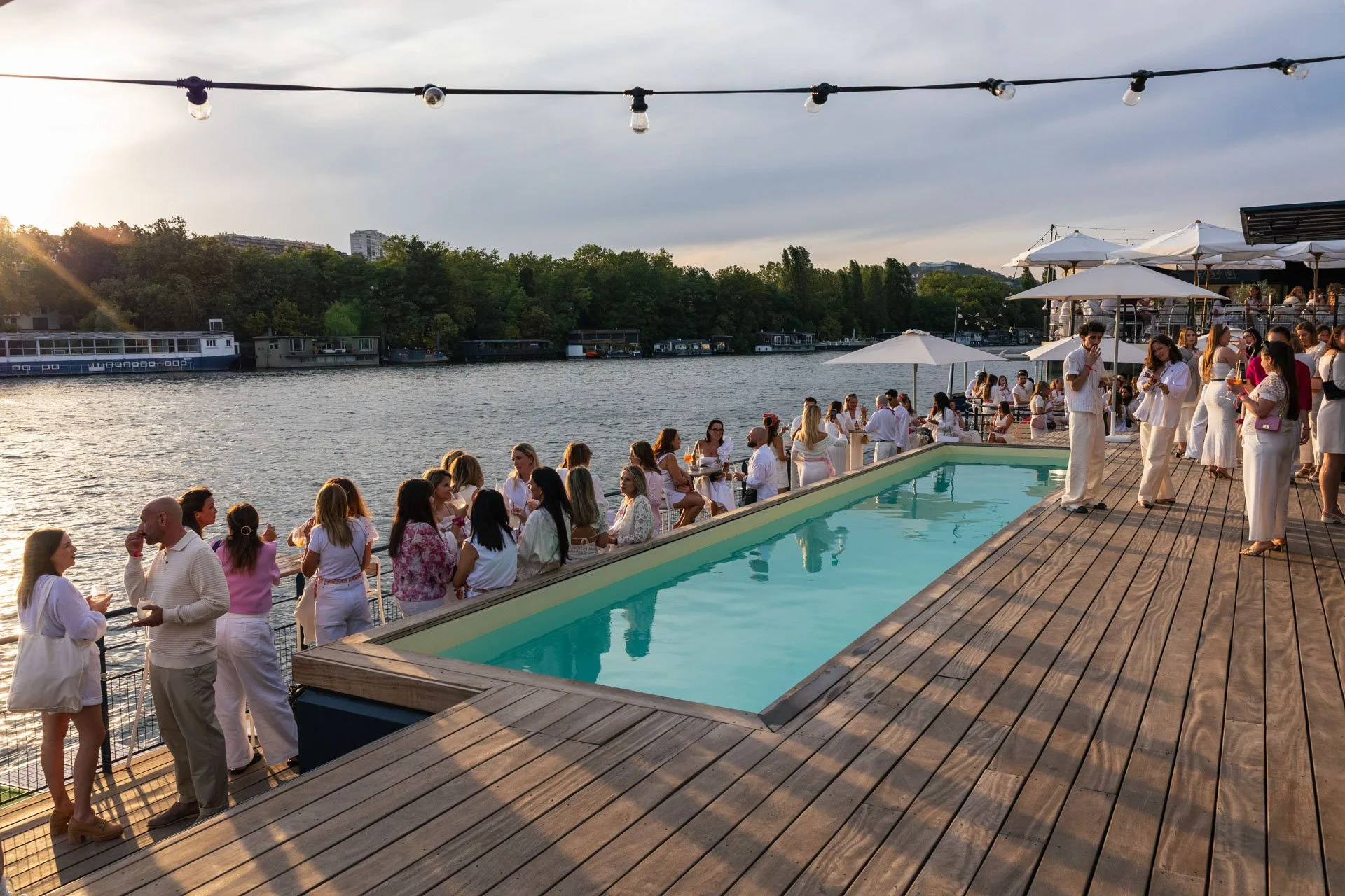 Une scène de fête en plein air au bord de l'eau avec beaucoup de personnes habillées en blanc, un espace piscine en bois, des parasols blancs, des lumières suspendues, et un environnement naturel avec des arbres et des bateaux sur la rivière en arriè