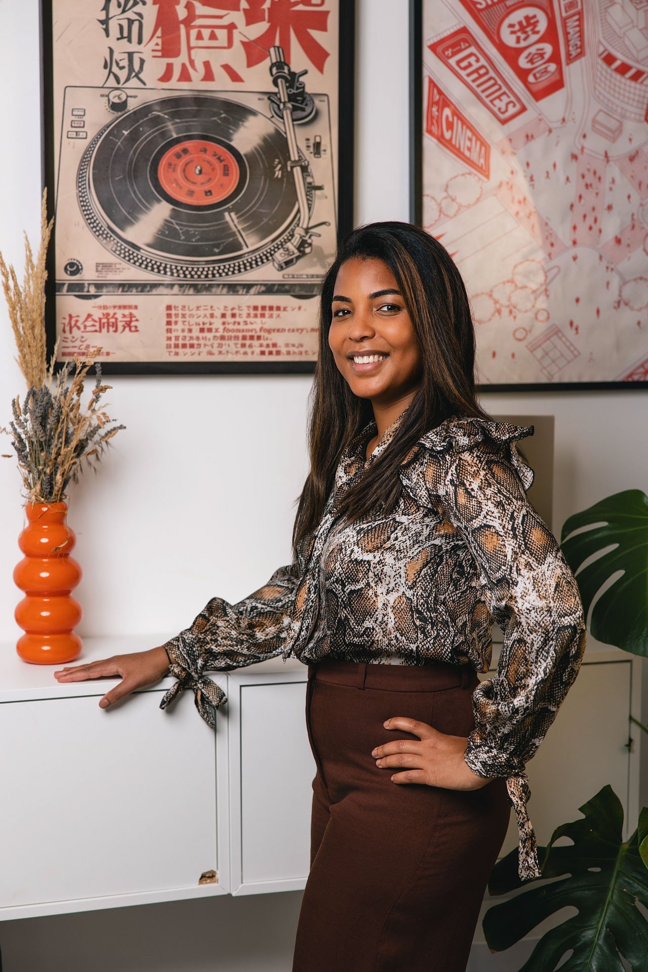 Une femme souriante pose dans un intérieur moderne, avec une étoffe d'imprimé serpent, une grande plante verte et des décorations murales, notamment une affiche de disque vinyle et une affiche avec du texte en japonais.