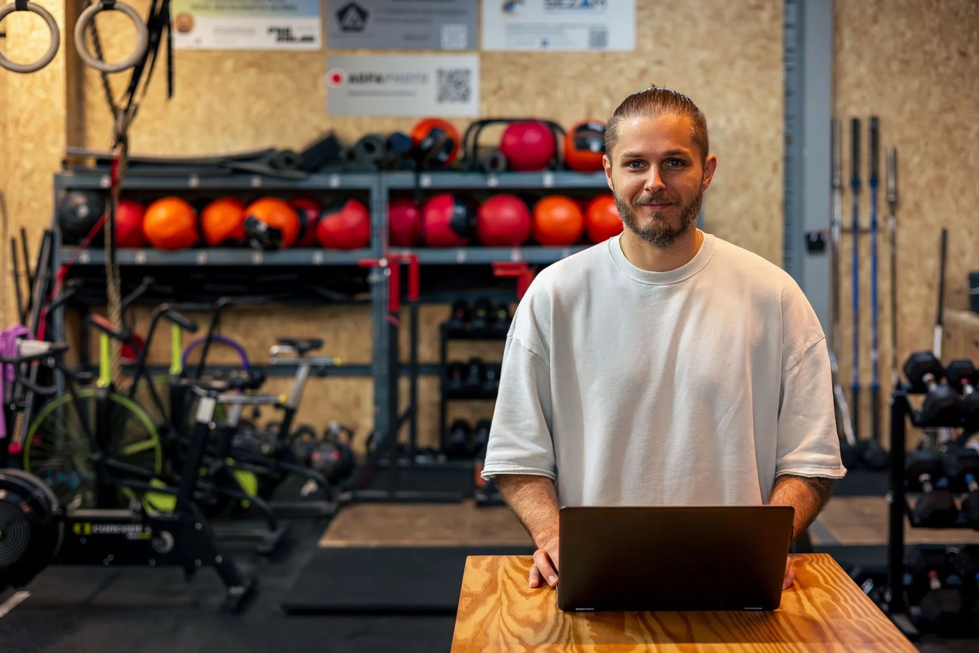 Un homme travaillant sur un ordinateur portable dans une salle de sport ou une salle d'entraînement avec des équipements de fitness, comme des balles, des vélos et des poids en arrière-plan.