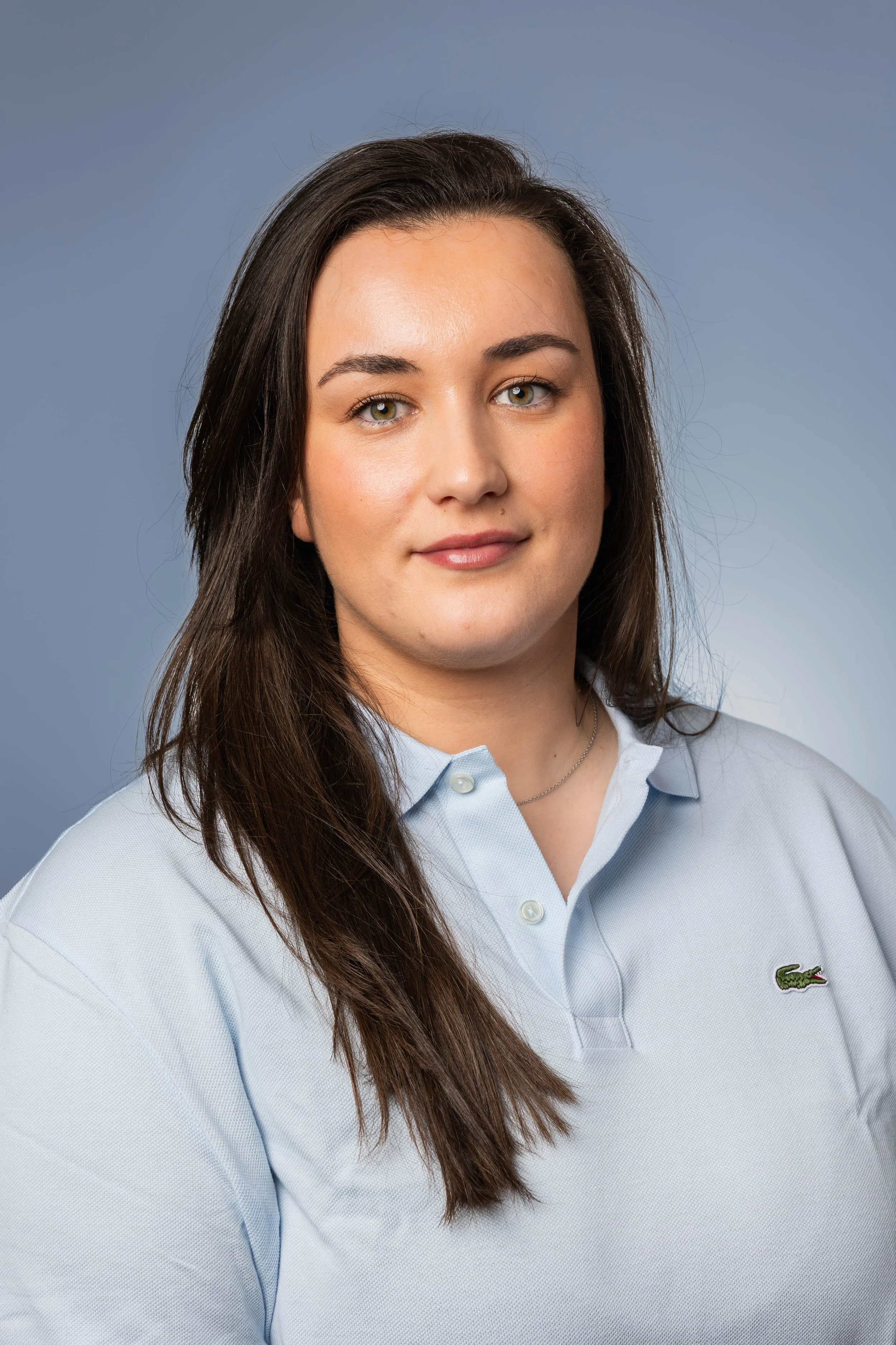 Une femme avec des cheveux longs bruns, portant une chemise bleue avec le logo Lacoste, sourit doucement devant un fond gris clair.