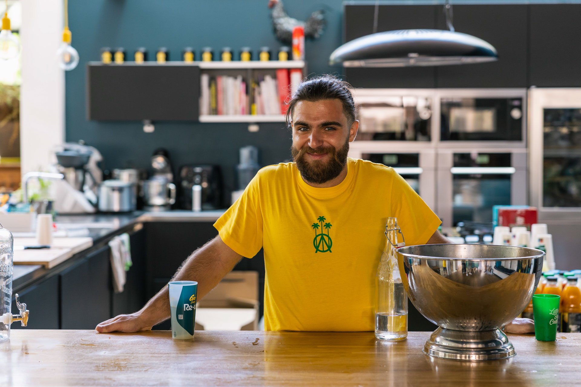 Un homme avec une barbe souriant dans une cuisine moderne portant un t-shirt jaune avec un logo de palmiers et de paix, devant un grand bol en acier inoxydable, deux verres d'eau, et plusieurs objets de cuisine.