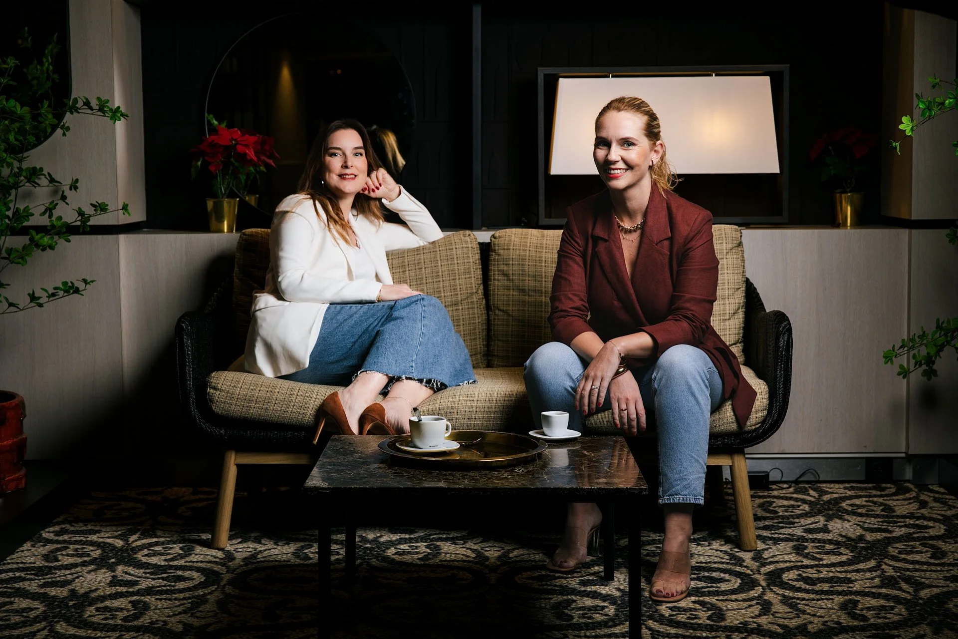 Deux femmes assises sur un canapé en bois et tissu beige, souriantes. La femme de gauche porte une veste blanche, une jupe en jean, et des talons marrons. La femme de droite porte une veste marron, un jean bleu clair, et des sandales. Il y a une tabl