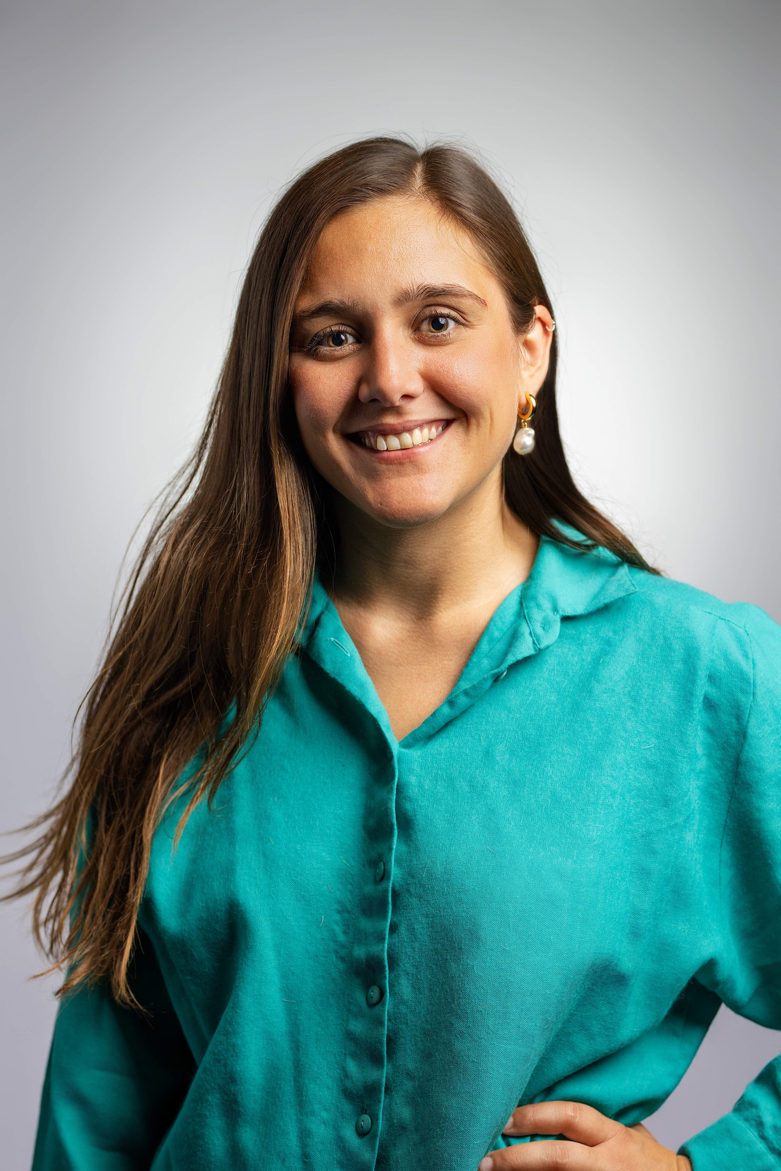 Jeune femme souriante avec cheveux longs, portant une chemise turquoise et une boucle d'oreille en perle, tenant sa taille avec la main gauche.