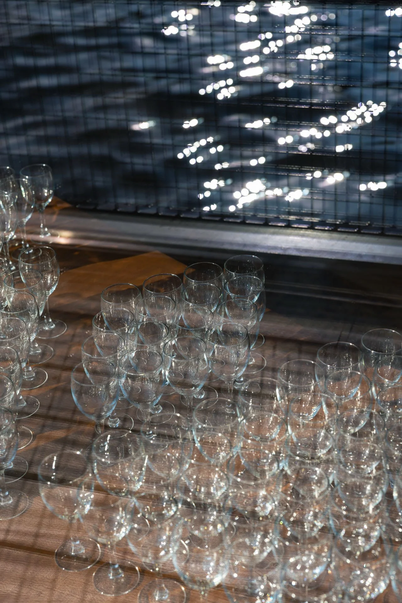 Plusieurs verres en cristal transparents sur une table en bois, avec des reflets de lumière sur une grille métallique en arrière-plan.