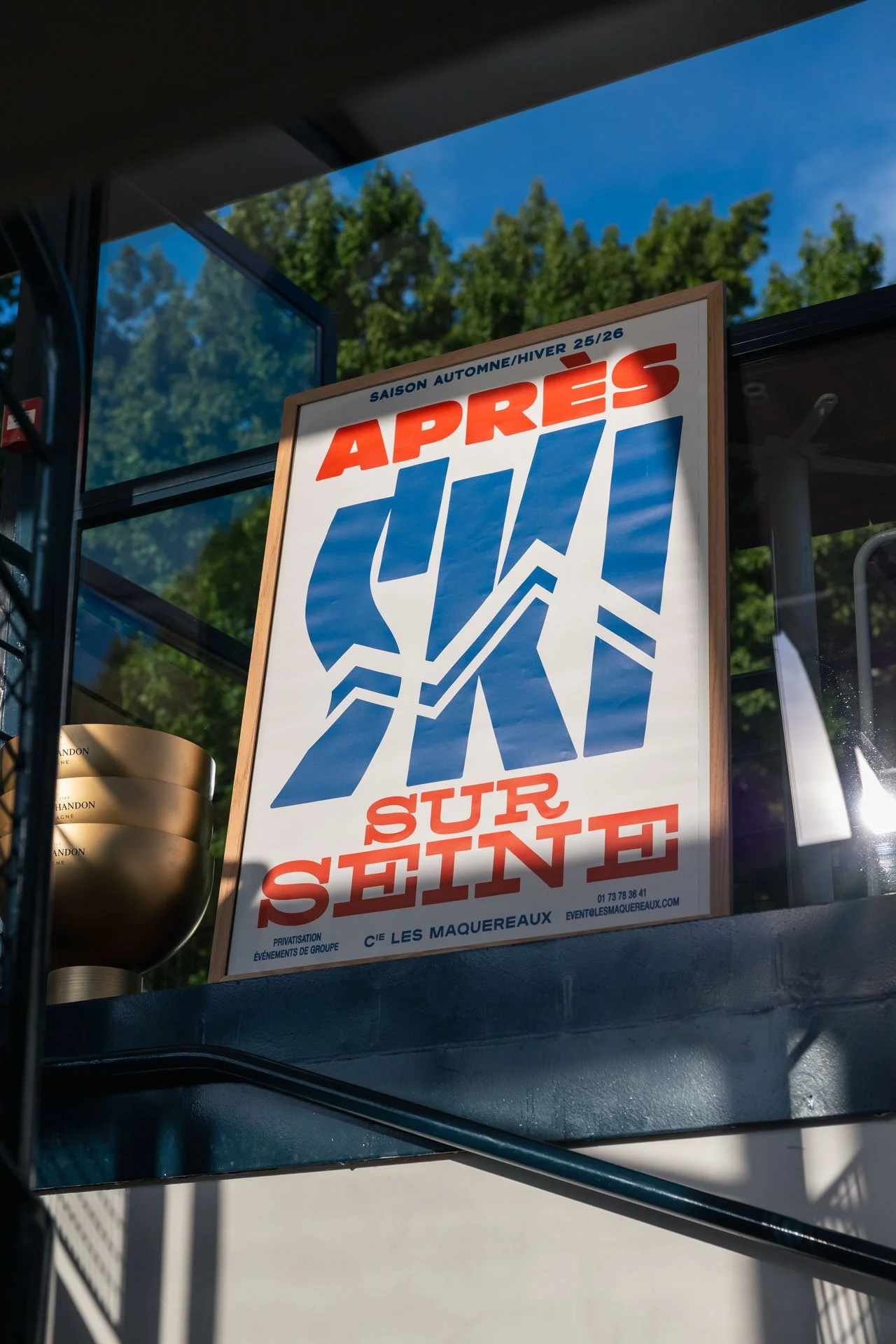 Affiche annonçant la sortie de la saison automne/hiver 25/26 avec le texte 'Après sur Seinde' et un logo bleu avec deux chevrons. Le panneau est placé dans un décor extérieur avec un ciel bleu et des arbres vertes en arrière-plan.