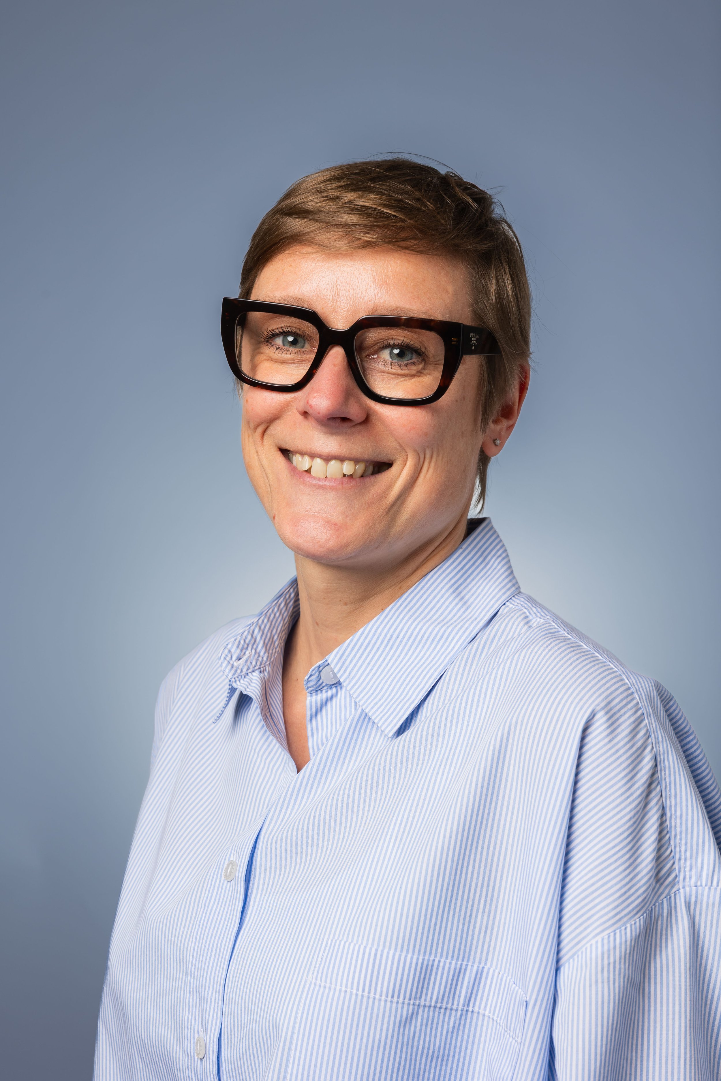 Une femme souriante portant de grosses lunettes à monture tortoise et une chemise bleue à rayures, sur un fond gris clair.