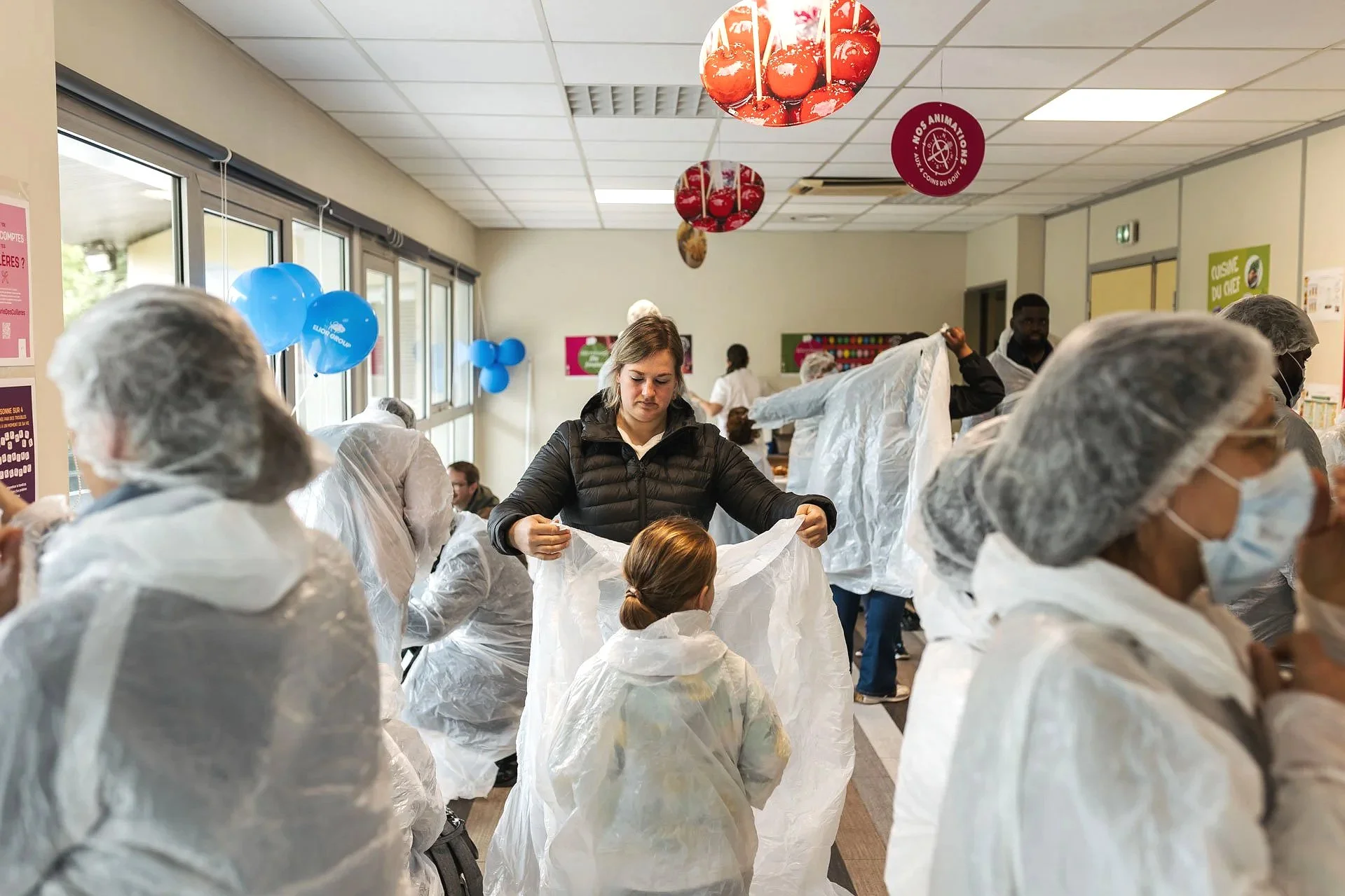 Des personnes portant des vêtements de protection blanche, collectives et diverses, participent à une activité en intérieur, dans une pièce à plafond suspendu. Certains individus ont des coiffes en filet ou en plastique, et il y a des ballons bleus a