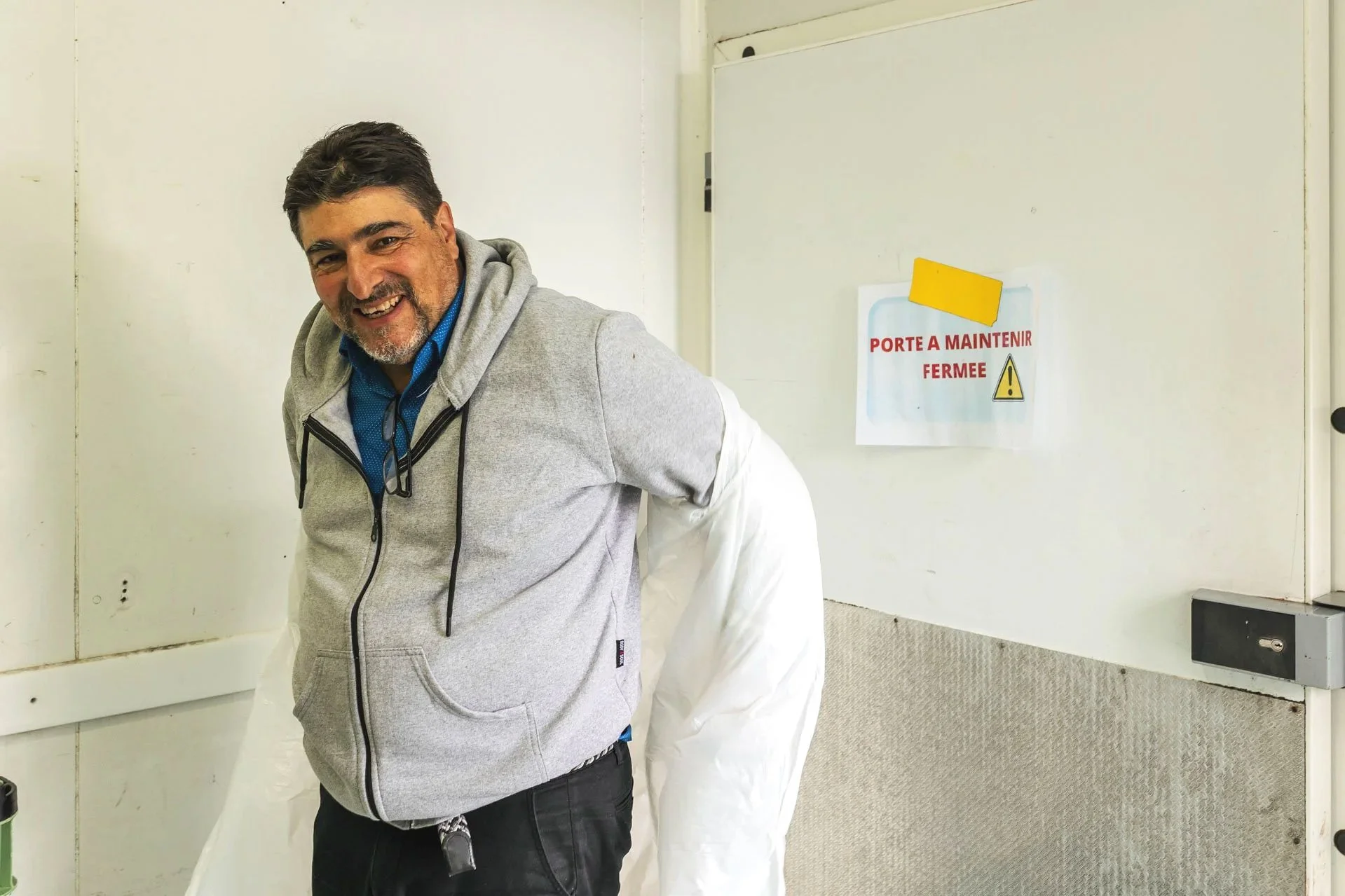 Homme souriant portant un sweat gris et une chemise bleue, dans une pièce avec un panneau blanc et un signe en français indiquant que la porte doit rester fermée.