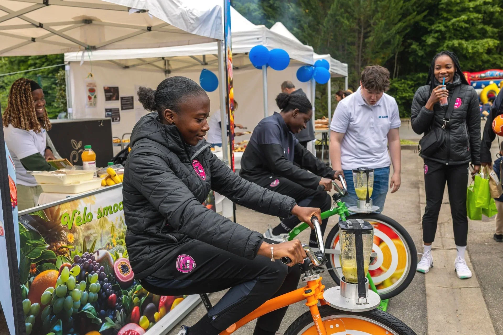 Groupe de personnes participant à une activité de dégustation ou d'animation en plein air, avec des tentes blanches, des ballons bleus, et une bannière avec des images de fruits.