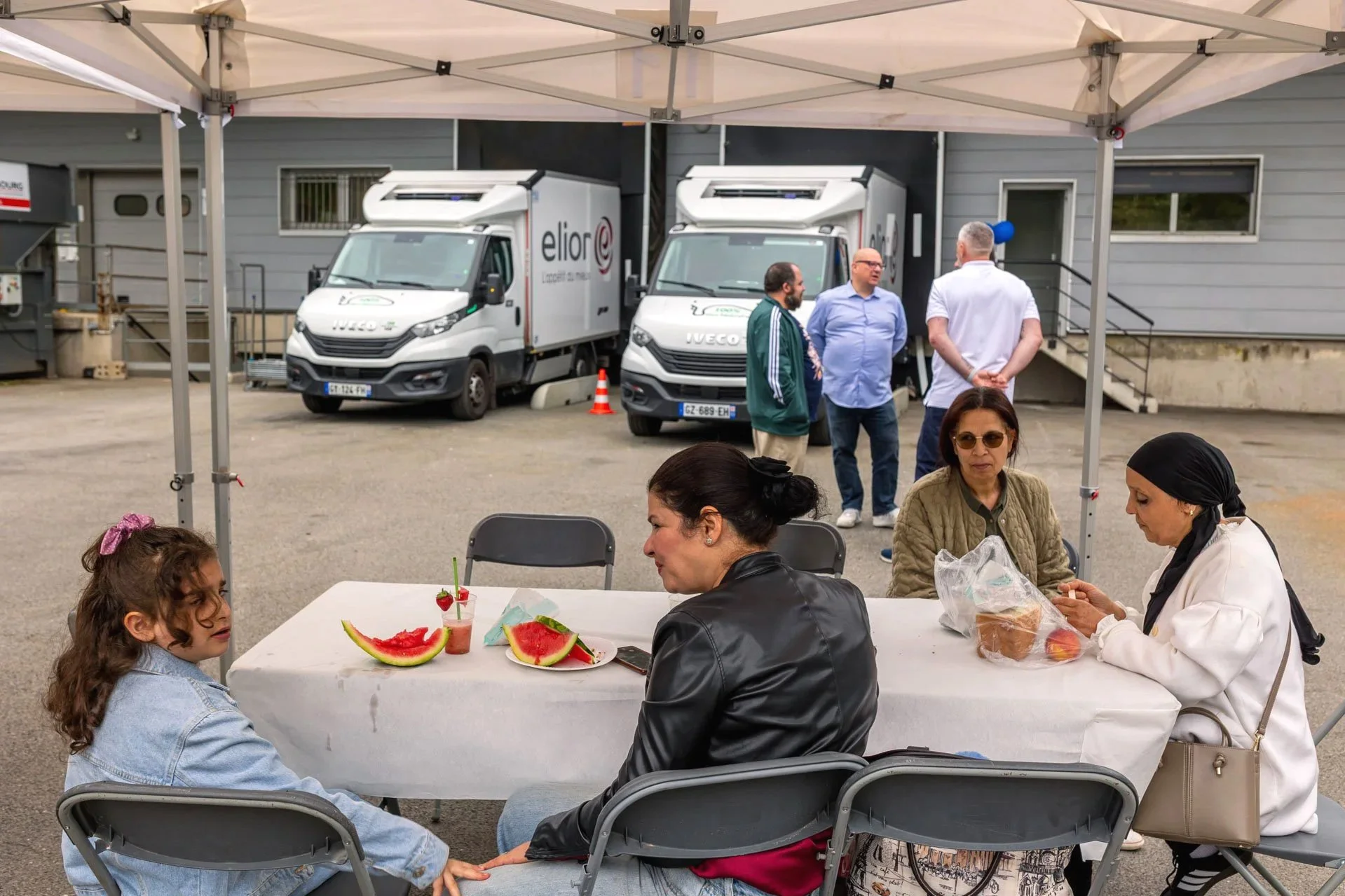 Groupe de personnes assises autour d'une table, partageant un repas avec des pastèques, sous une tente, avec deux camionnettes blanches en arrière-plan.