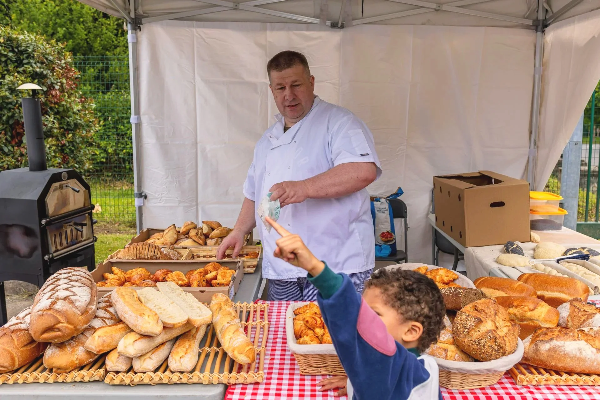 Un vendeur de boulangerie dans une tente en plein air vend du pain et des viennoiseries. Deux enfants sont visibles en train de choisir des produits.