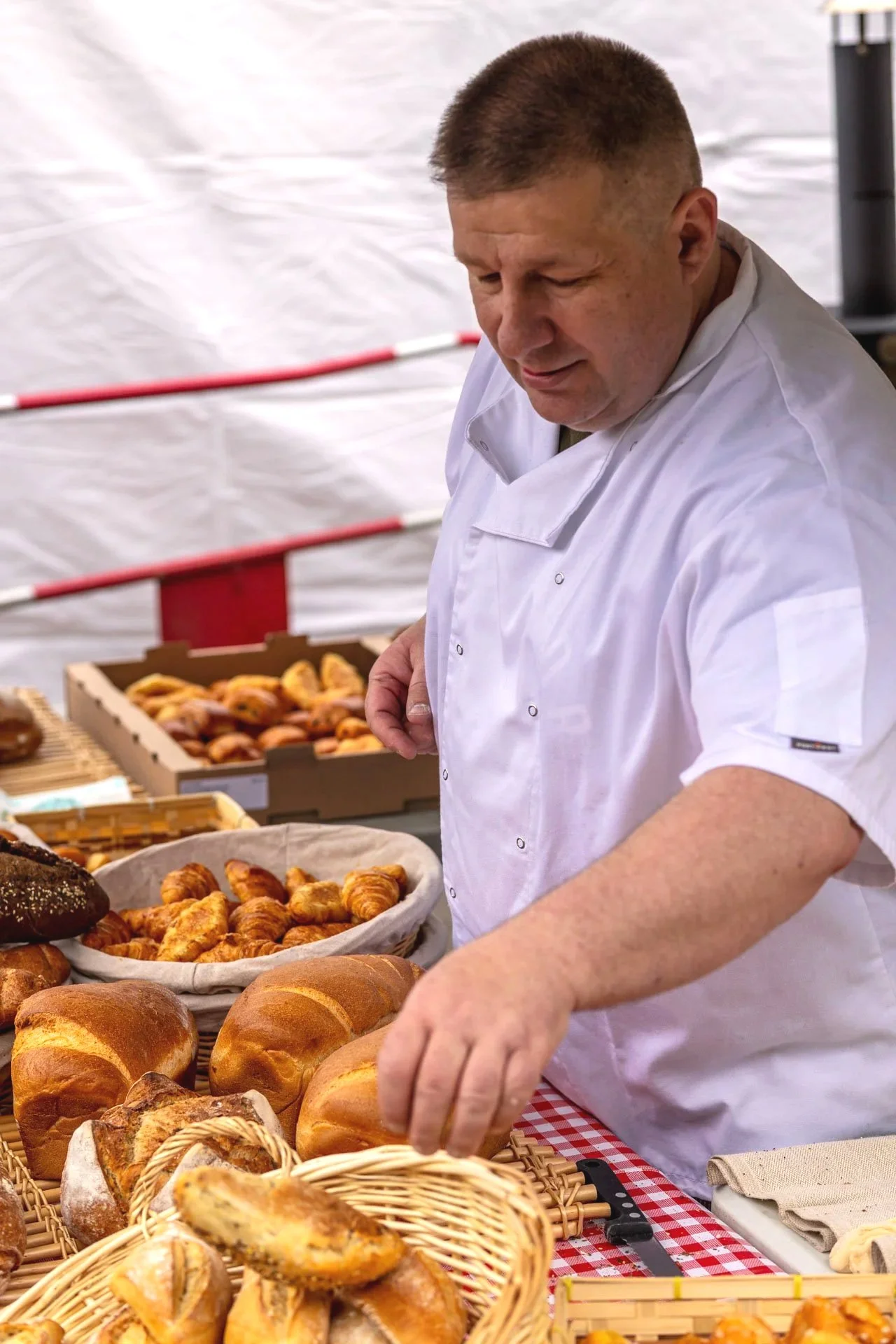Un homme avec une veste blanche vendant des pâtisseries dans un marché. Il y a des croissants, pains, et autres viennoiseries sur la table. En arrière-plan, des étagères avec plus de pâtisseries.