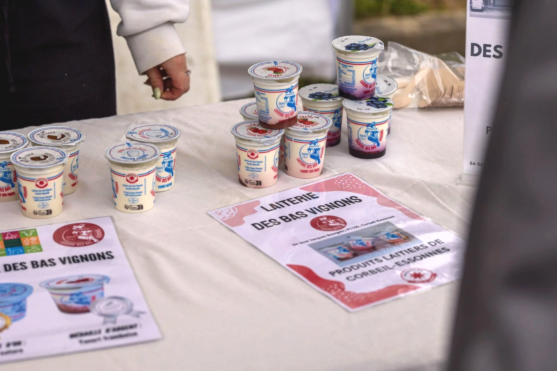 Des pots de yaourt à la fraise et aux fruits déposés sur une table, avec des affiches qui mentionnent la laiterie des Bas Vignons.