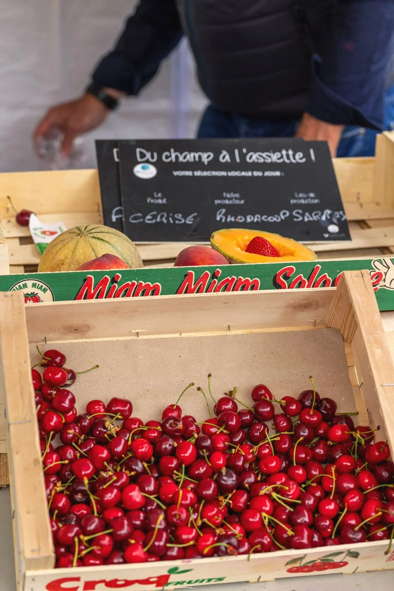 Une caisse en bois remplie de cerises rouges, avec une pastèque et des pêches en arrière-plan. Un tableau noir indique la sélection locale du jour, comprenant la cerise, la pêche, la framboise et la fraise.