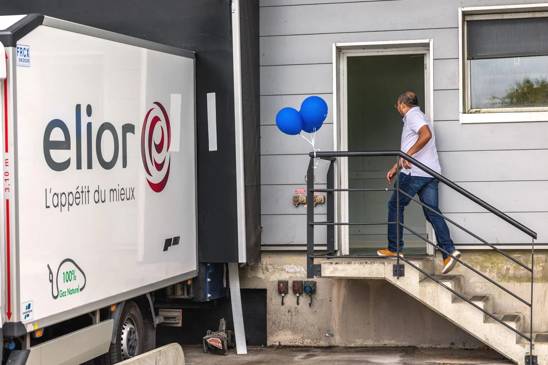 Une personne descend un escalier extérieur devant un bâtiment gris, à côté d'un camion blanc avec un logo et le slogan 'L'appétit du mieux'.