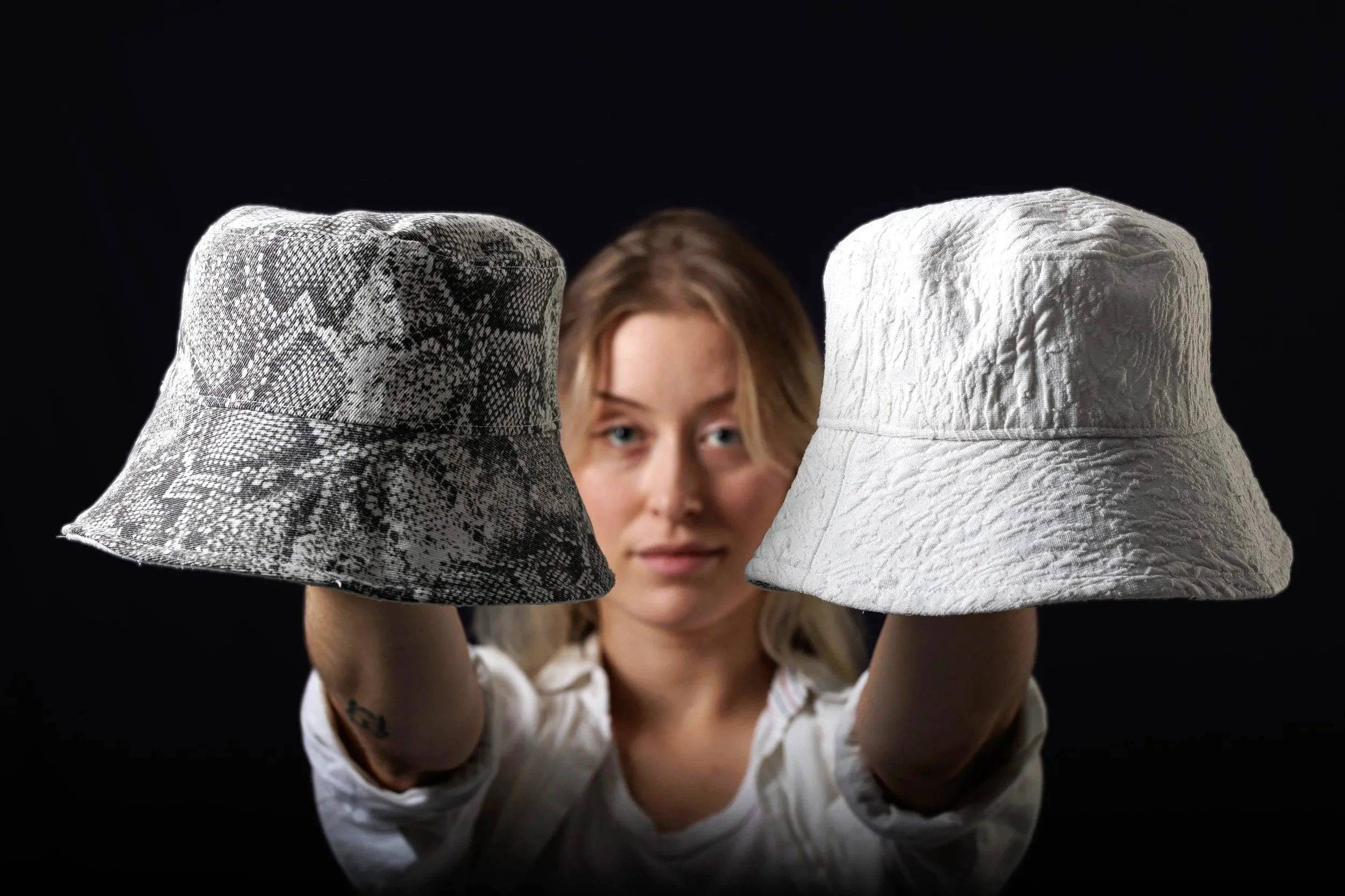 Une jeune femme regarde directement l'objectif tout en tenant deux chapeaux, l'un à motif serpent et l'autre en tissu blanc, devant un fond noir.