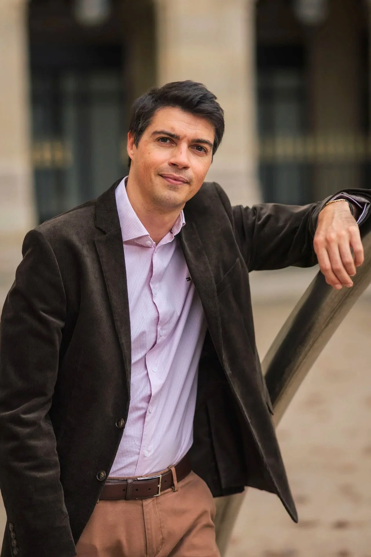 Un homme en costume décontracté pose avec un sourire devant un bâtiment. Il porte une chemise rose clair, une veste marron et un pantalon beige, avec une ceinture en cuir.