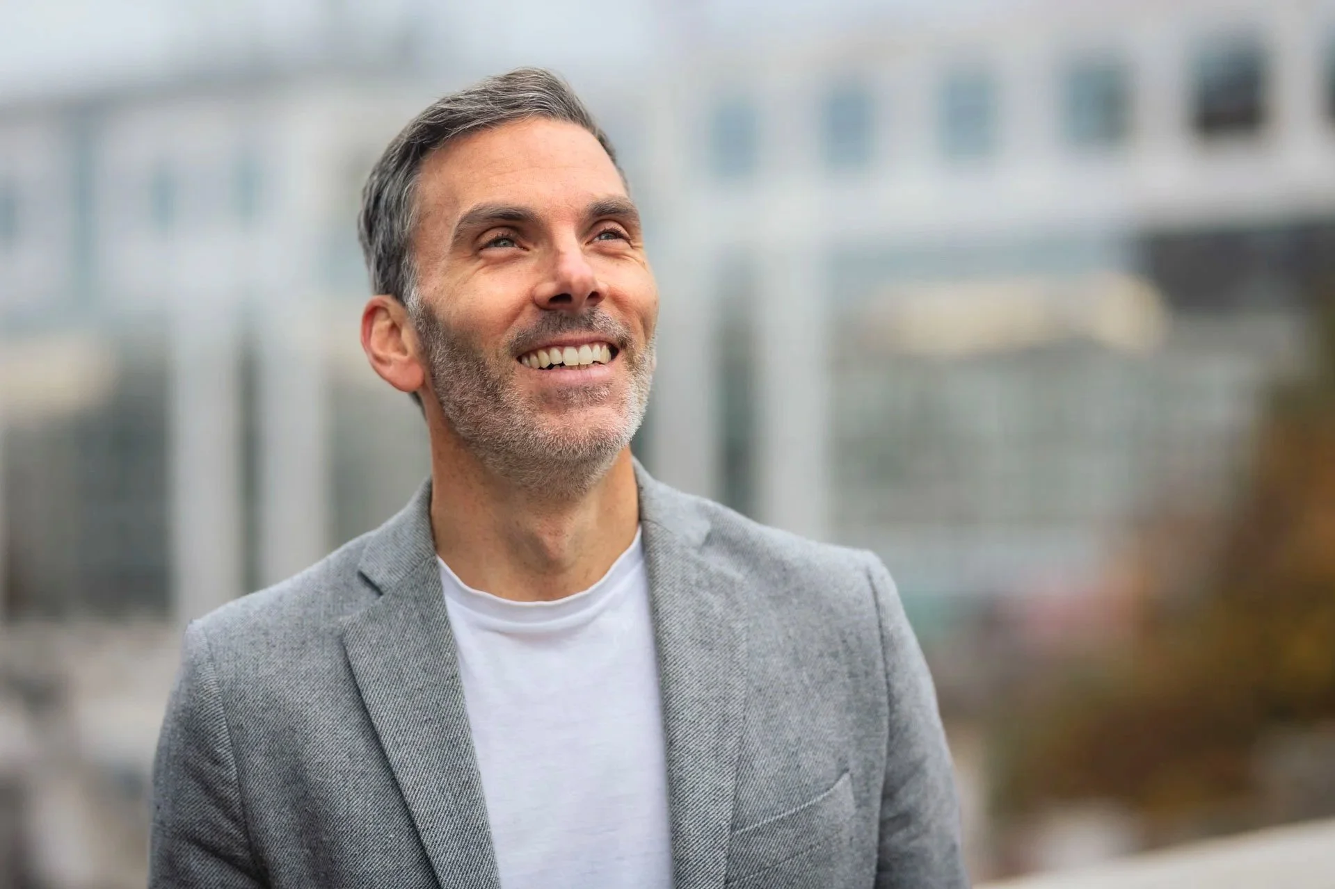 Un homme souriant portant un costume gris clair et une chemise blanche, avec un arrière-plan flou d'un bâtiment moderne.