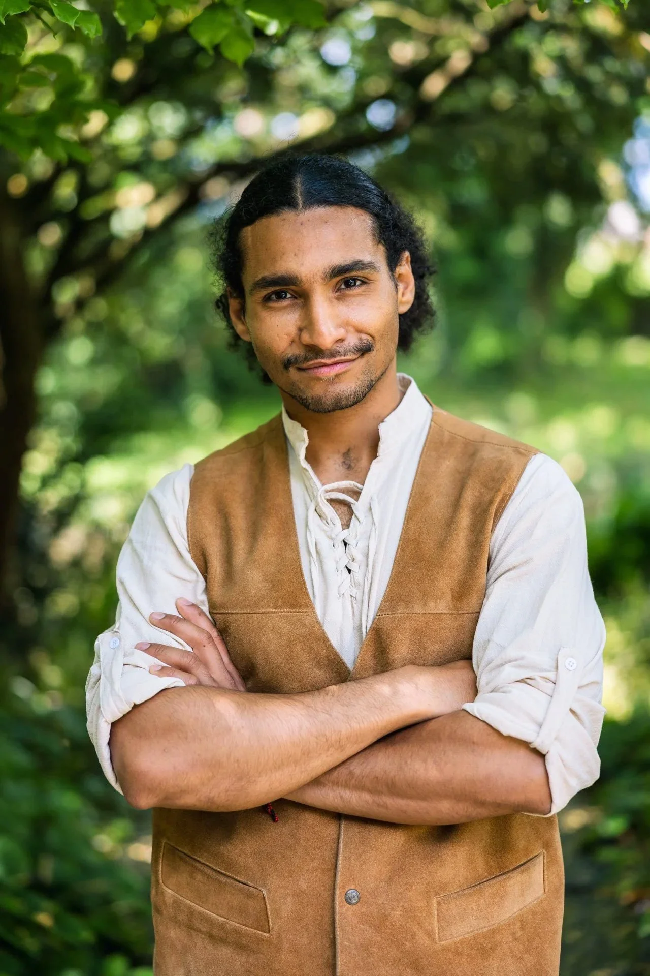 Un homme d'une peau sombre porte une chemise écrue et un gilet en daim marron, croisant les bras, devant un fond de feuillage vert et lumineux dans la nature.