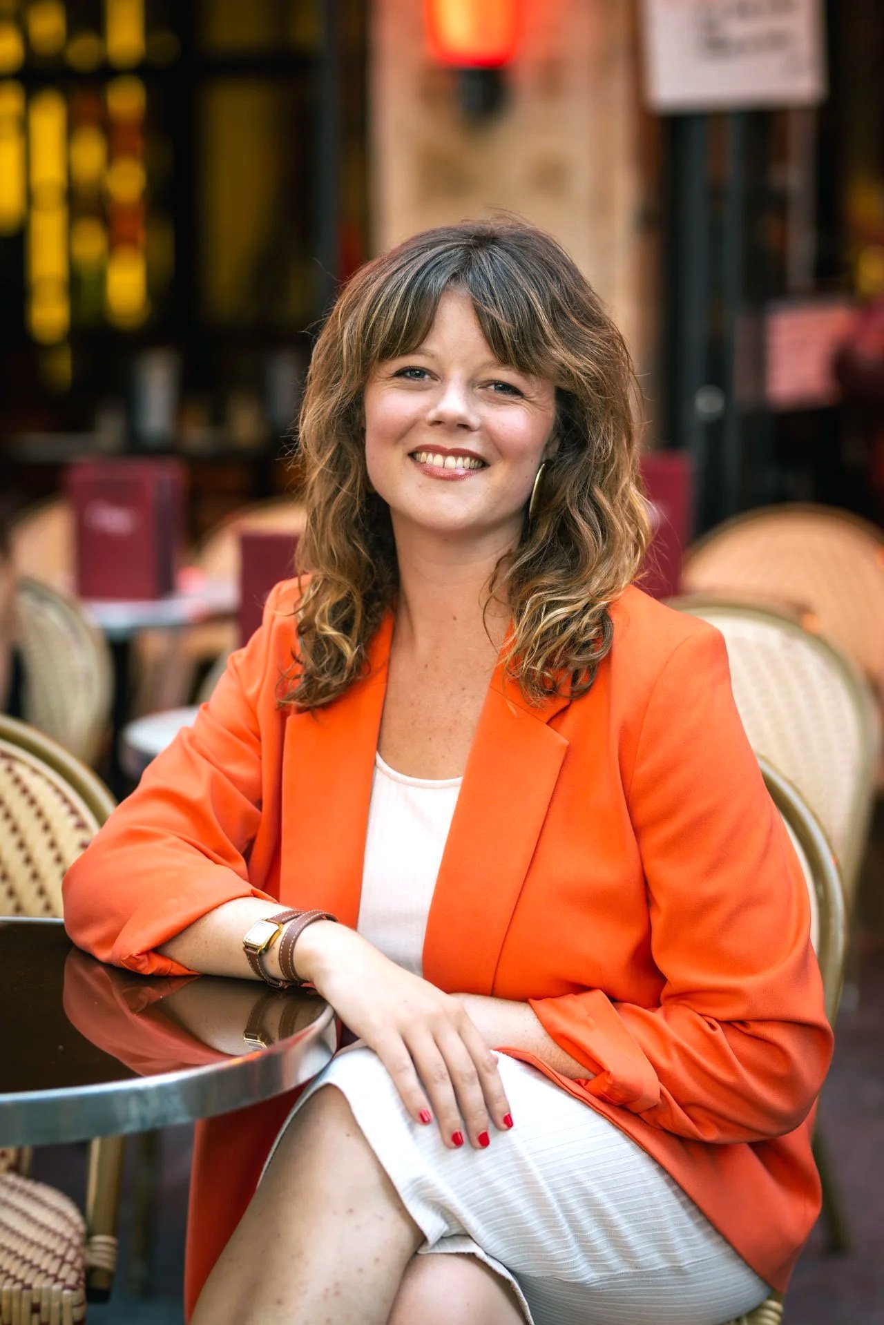 Femme souriante portant une veste orange, assise à une table dans un restaurant, avec des chaises en rotin autour.