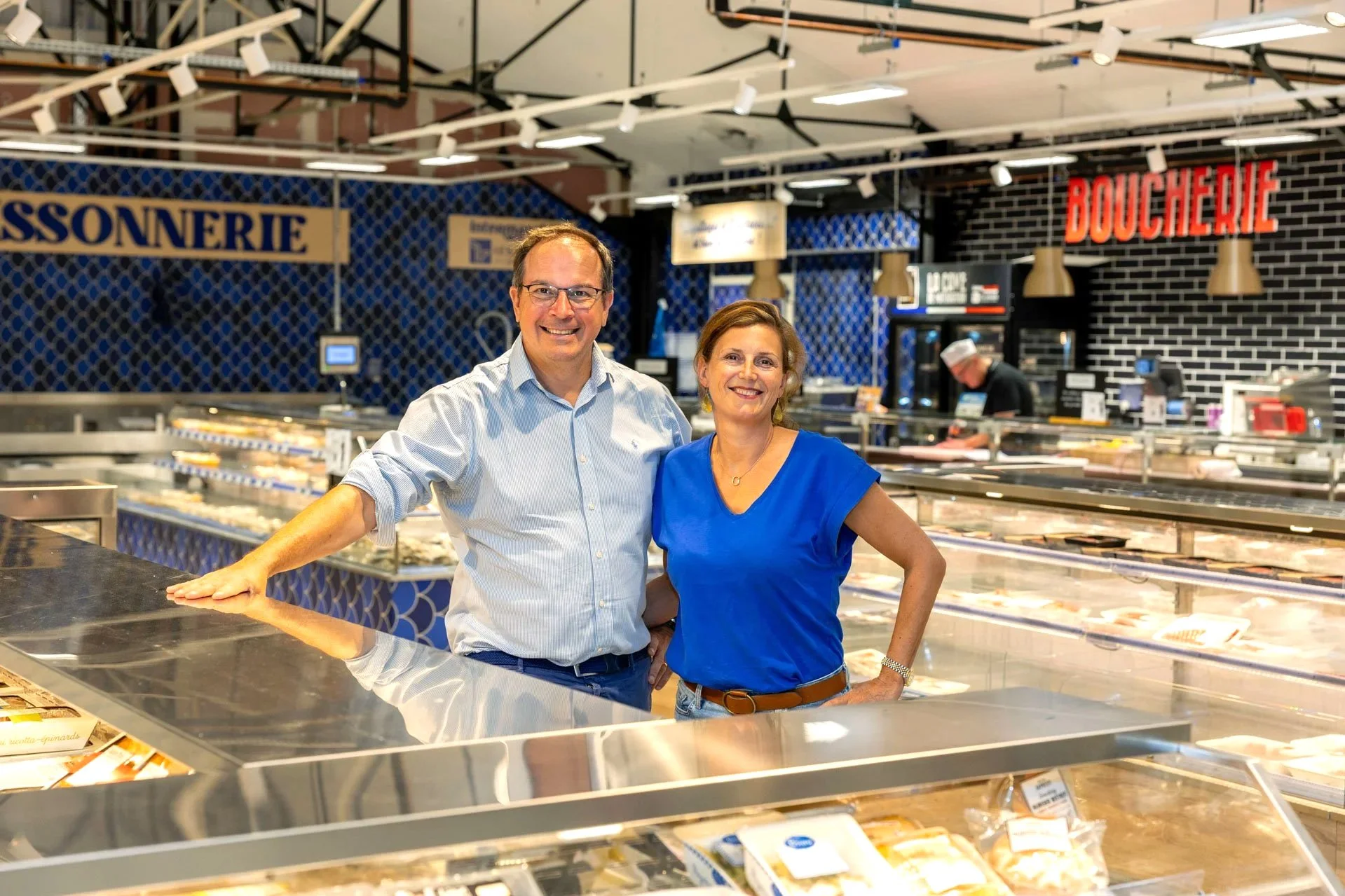 Deux personnes, un homme et une femme, posent dans une boucherie ou un rayon de viande, souriant à la caméra.