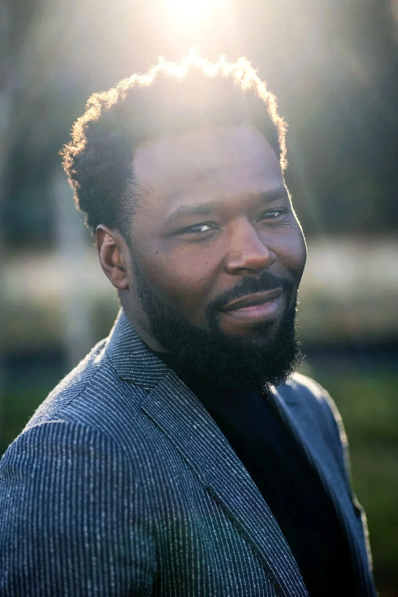 Un homme souriant avec une barbe, portant une veste à rayures, en extérieur avec un rayonnement solaire en arrière-plan.
