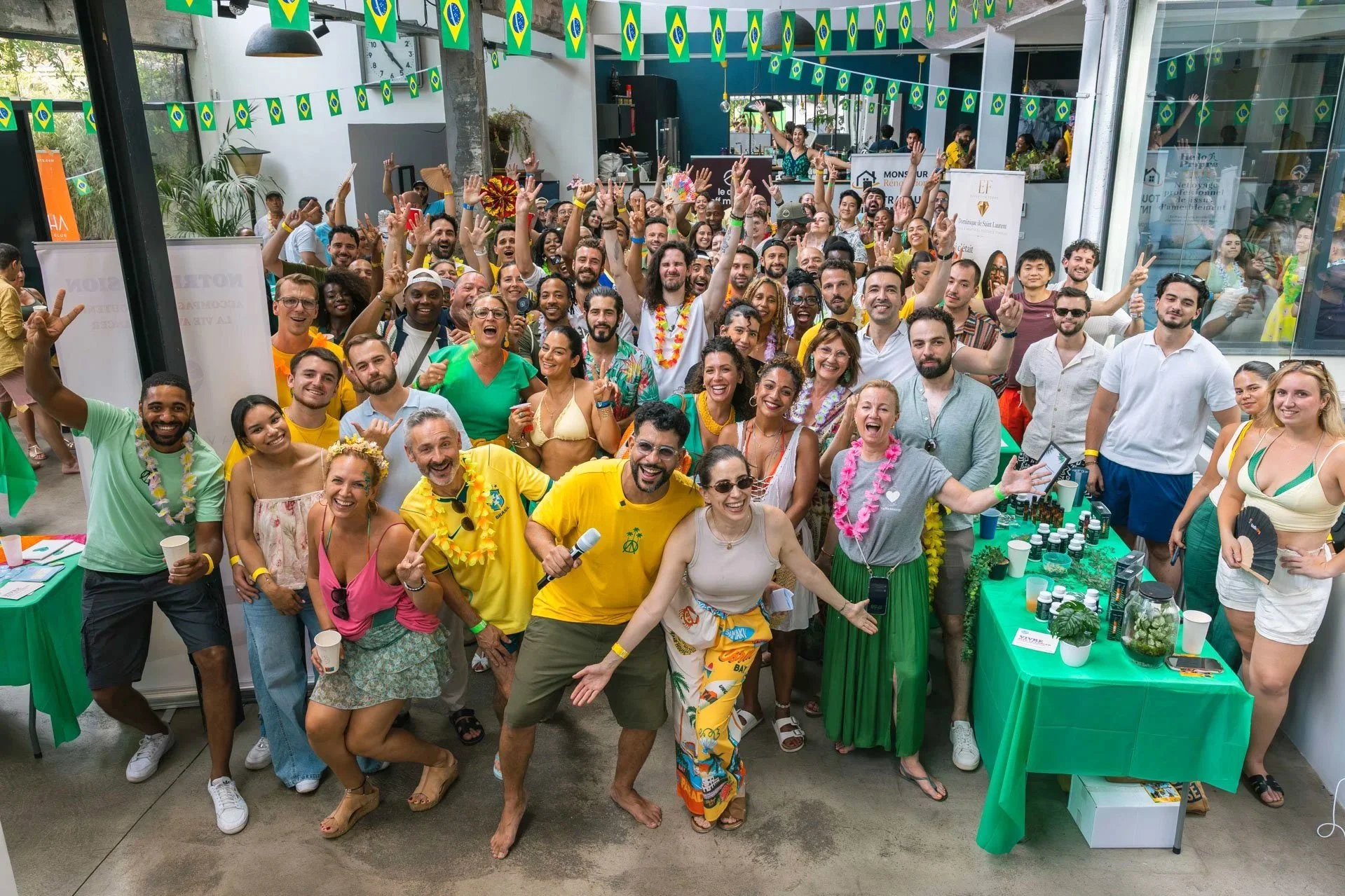 Groupe de personnes riant et posant pour une photo lors d'une fête ou événement tropical avec décor vert et drapeaux du Brésil suspendus au plafond.