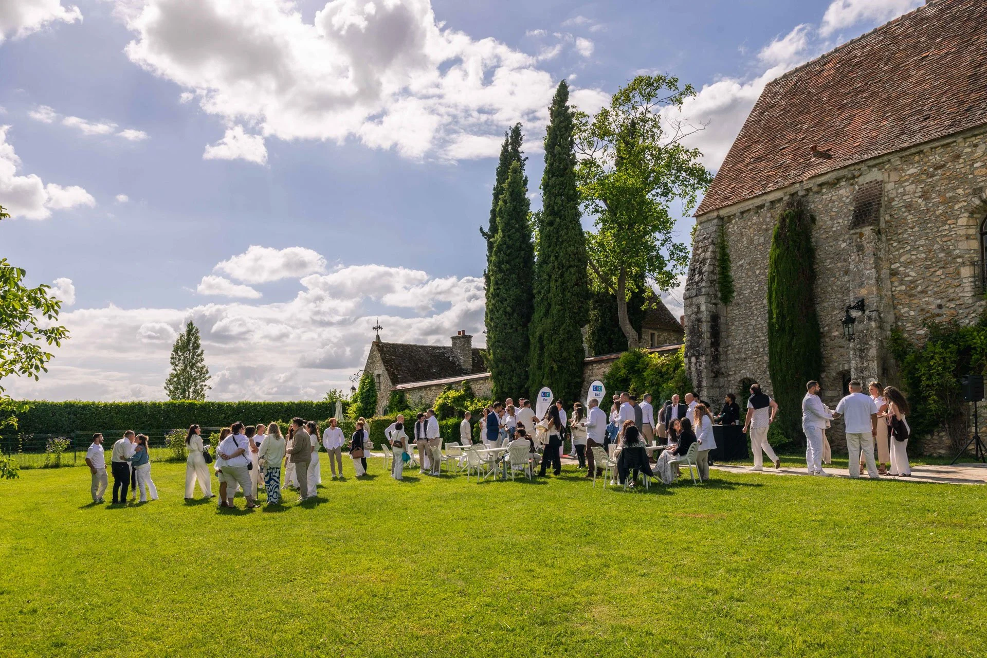 Groupe de personnes en habit formel lors d'une réception en plein air devant une vieille église en pierre, avec un ciel partiellement nuageux.
