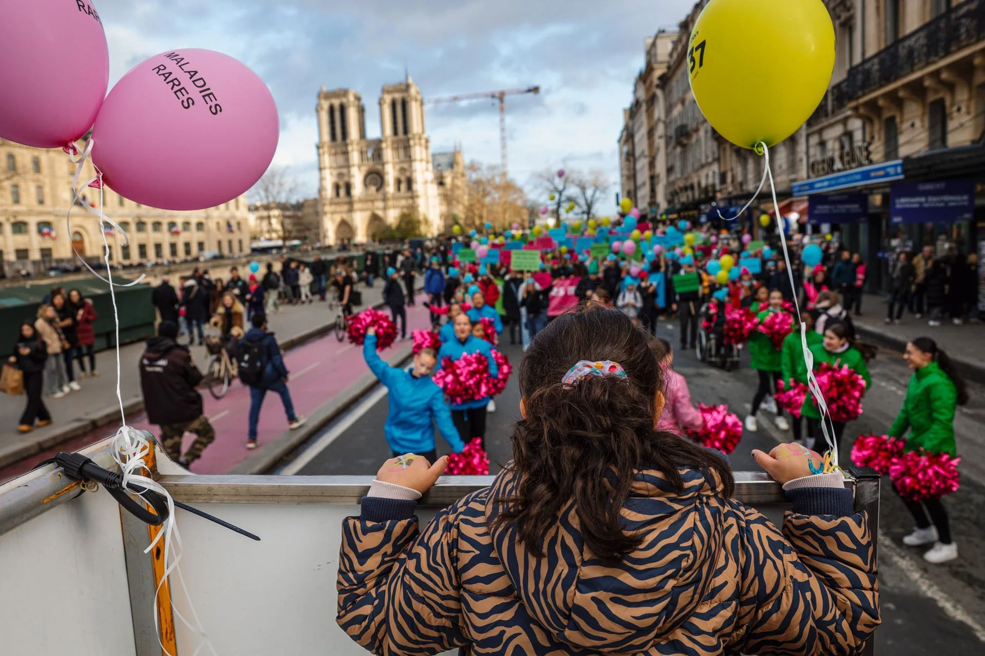 Fête dans les rues de Paris avec de nombreuses personnes, ballons colorés, et des marcheurs. Vue partielle d'une jeune fille regardant la parade depuis une camionnette, avec la cathédrale Notre-Dame en arrière-plan.
